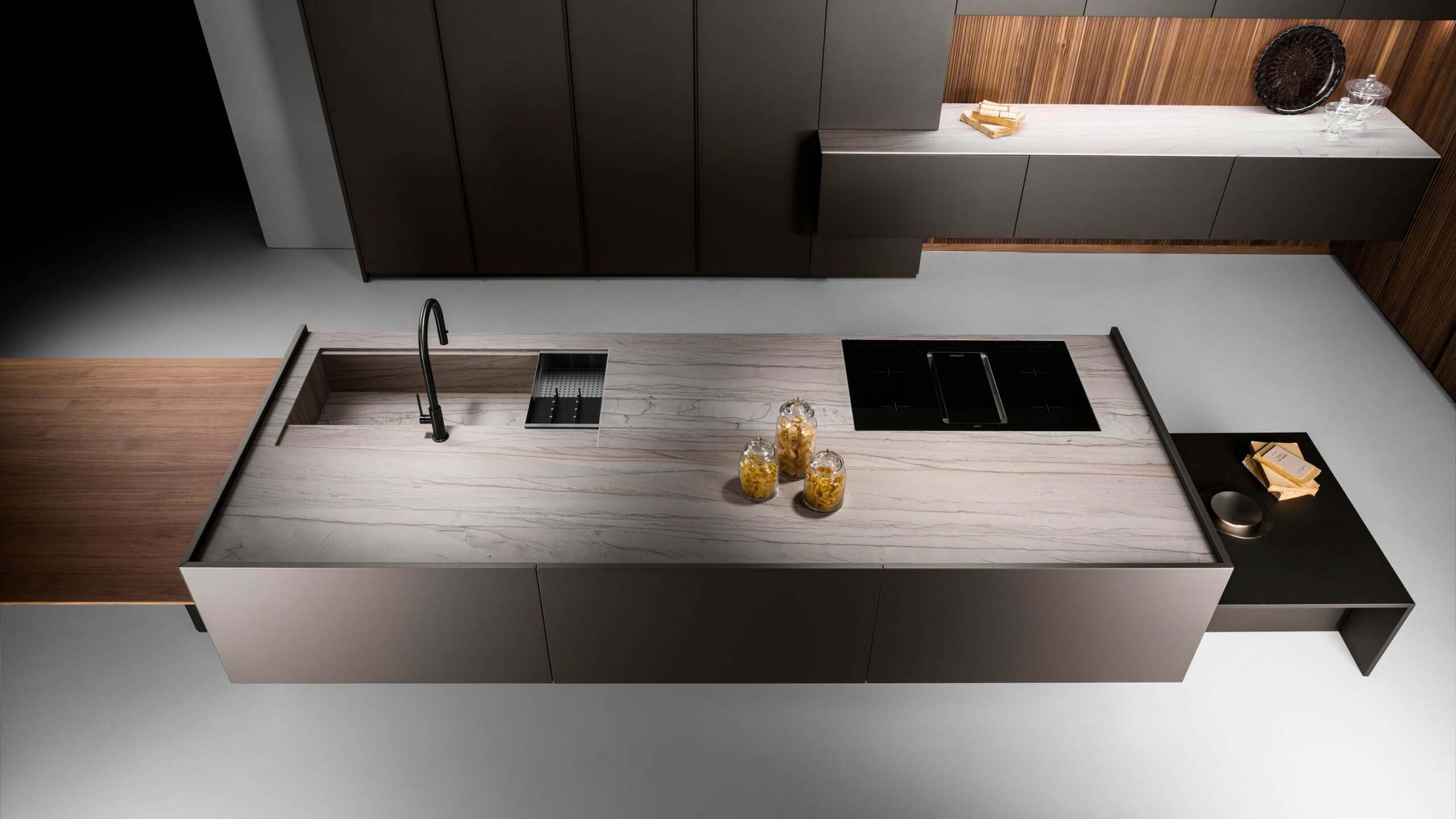 An angled shot of a high-end minimalist kitchen and dining area. The kitchen features a large, light-colored island with an integrated hob and a dedicated seating area. The back wall is covered in luxurious stripped walnut oak veneer, and a black, cylindrical extractor hood hangs above the island.