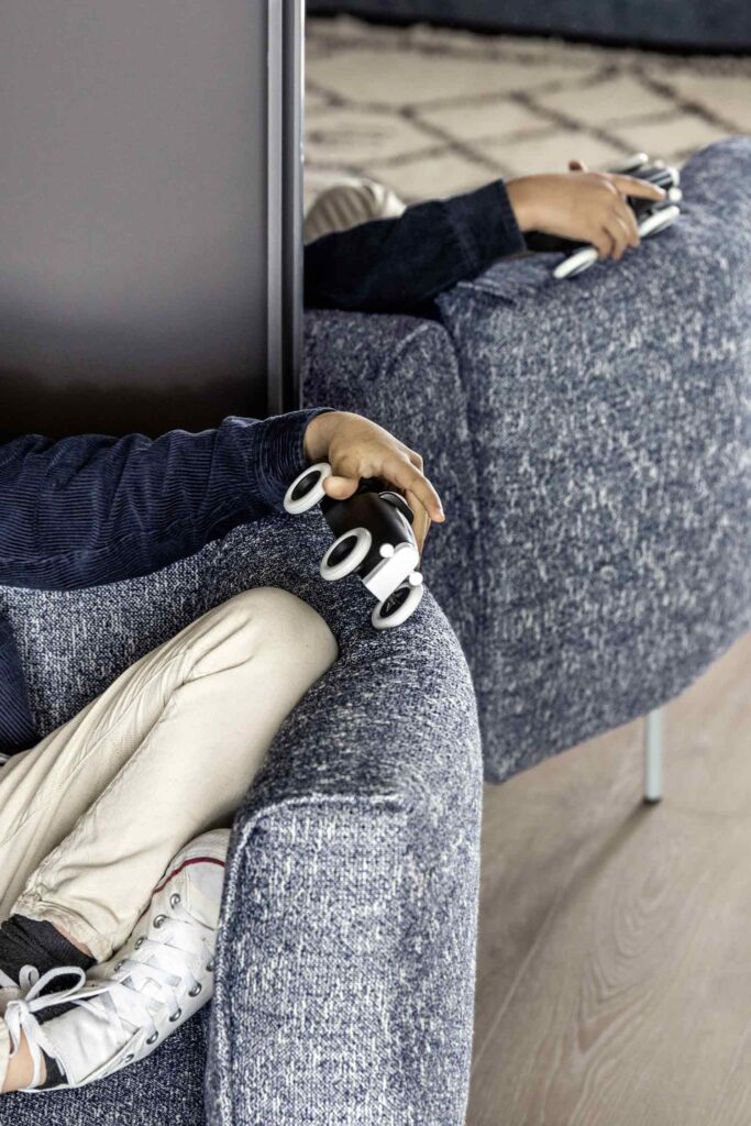 A cropped, high-angle shot of a person relaxing in a blue and white woven fabric armchair. The person's hand rests on the armrest, holding a small black flashlight with a white base. Their feet, wearing off-white pants and white sneakers, are visible in the foreground. Another person is partially visible in a matching armchair behind them, holding a similar item. The chairs have thin metal legs and are separated by a gray partition.