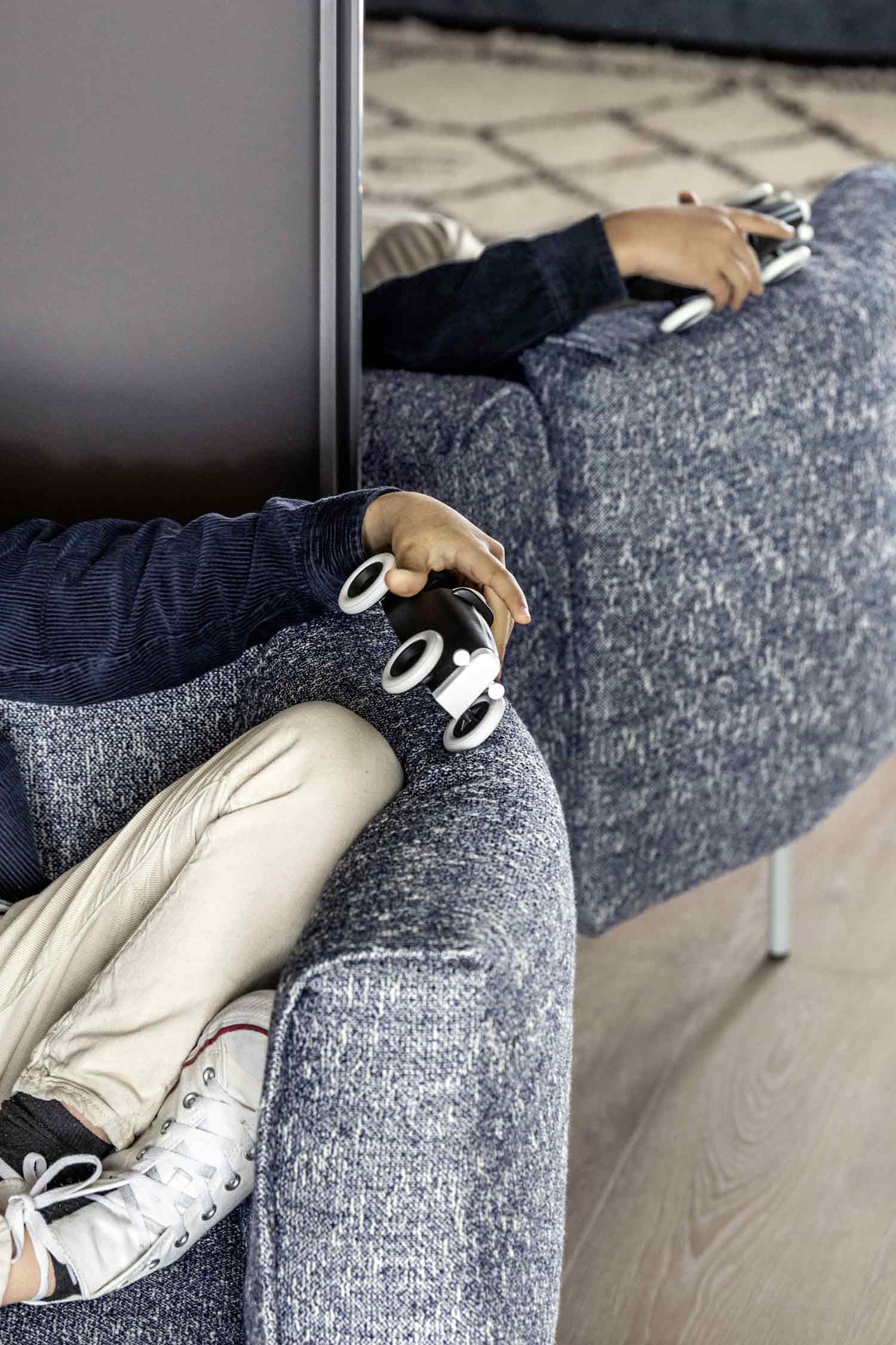 A cropped, high-angle shot of a person relaxing in a blue and white woven fabric armchair. The person's hand rests on the armrest, holding a small black flashlight with a white base. Their feet, wearing off-white pants and white sneakers, are visible in the foreground. Another person is partially visible in a matching armchair behind them, holding a similar item. The chairs have thin metal legs and are separated by a gray partition.
