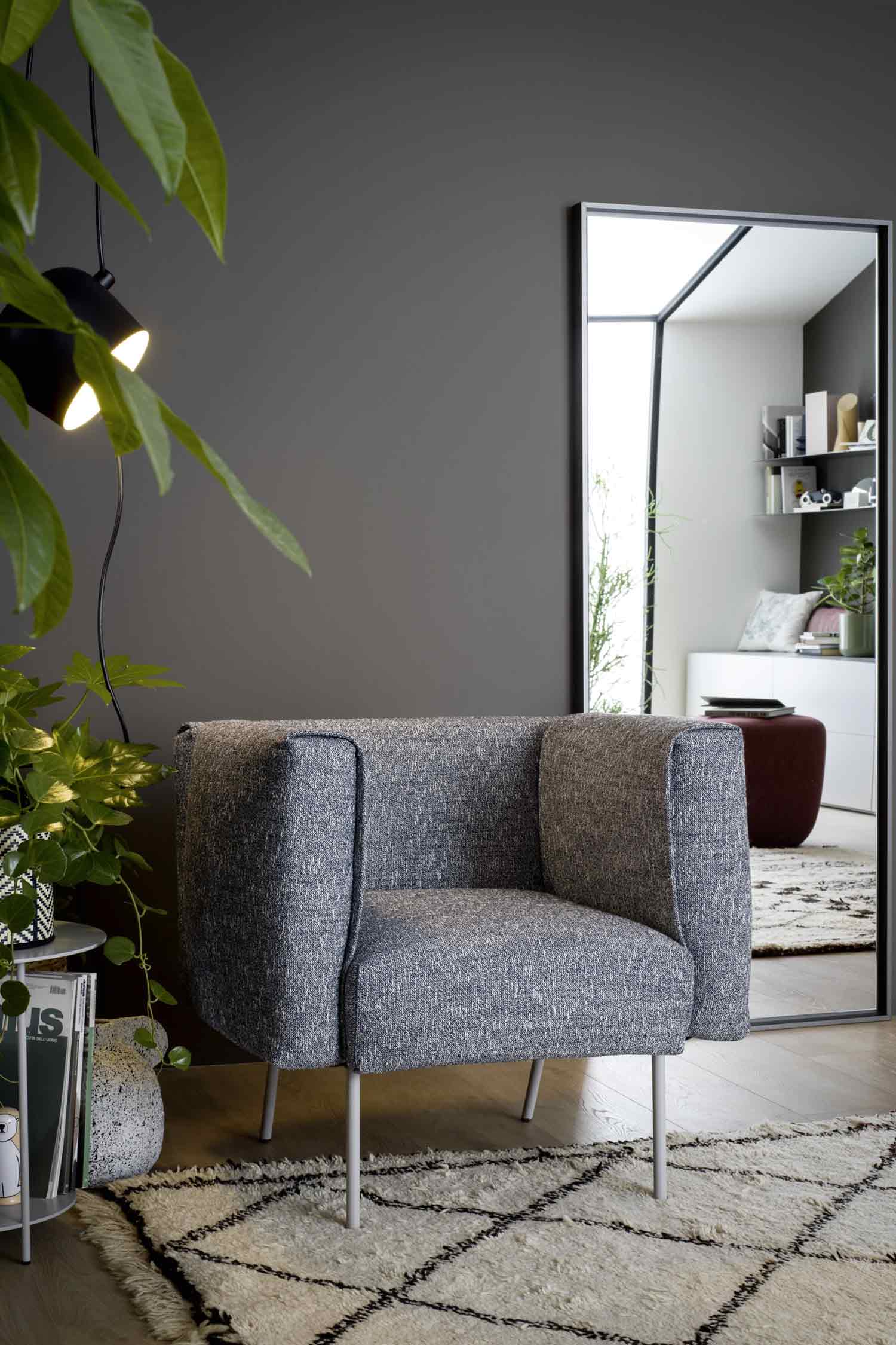 A cozy gray and white woven fabric armchair with thin light gray metal legs sits on a white shag rug with a black geometric pattern. To the left of the chair, a leafy green plant hangs down in front of two modern black lights. To the right, a large, dark-framed mirror leans against a dark gray wall, reflecting a modern white interior with a red ottoman and white shelves.
