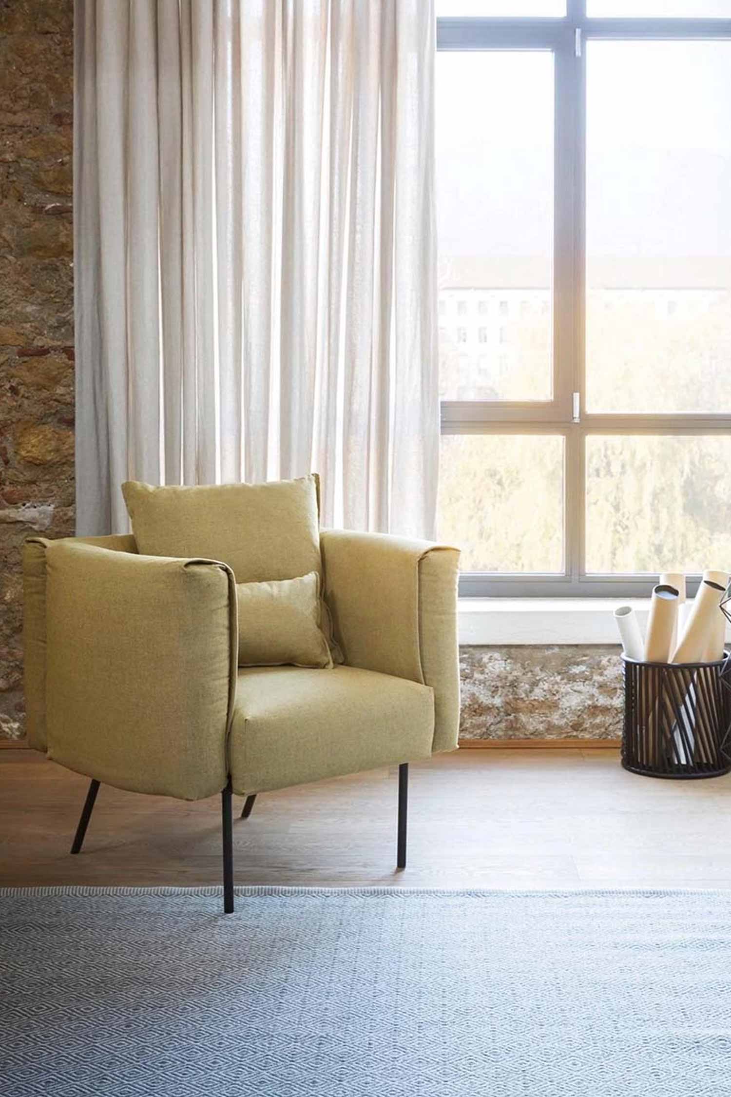 A plush armchair with an olive green fabric and a lumbar pillow sits on a light wood floor. The chair has thin black metal legs. A light gray rug with a subtle chevron pattern is in the foreground. To the right of the chair, a black wire basket holds rolled-up white paper. The background features a large window with sheer white curtains on the left and a stone wall on the right.
