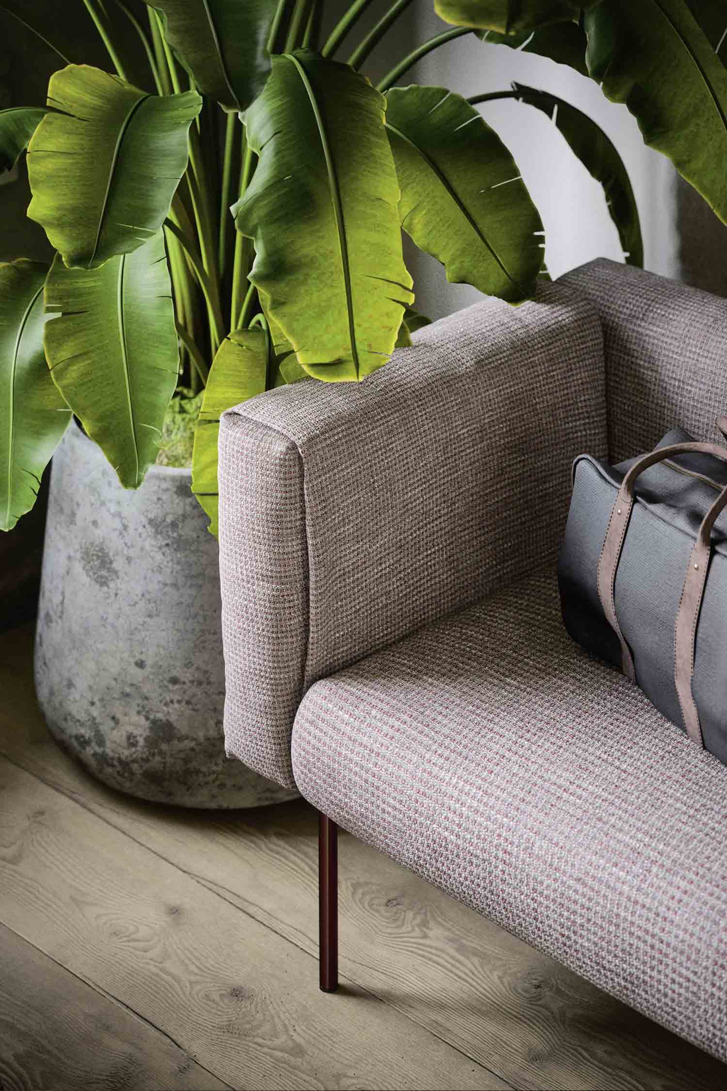 A close-up of a modern sofa with textured gray and pink fabric, a large leafy plant in a concrete pot, and a brown leather bag on the sofa.