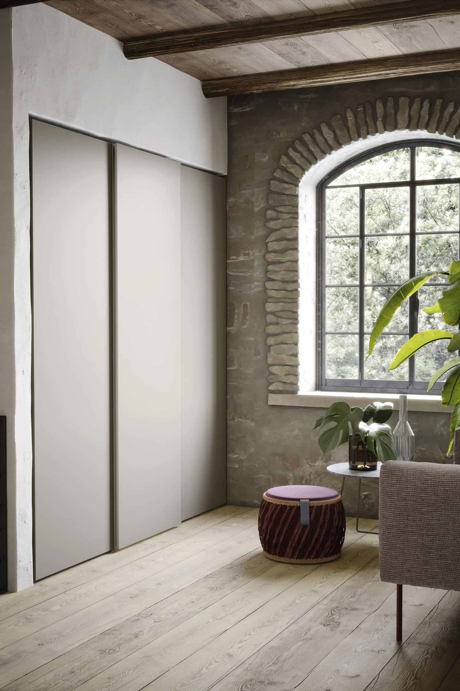 A bright, rustic-style room with a large arched window and exposed brick. The room features a light wood floor, modern sliding closet doors, a small round table with plants, and a woven ottoman.