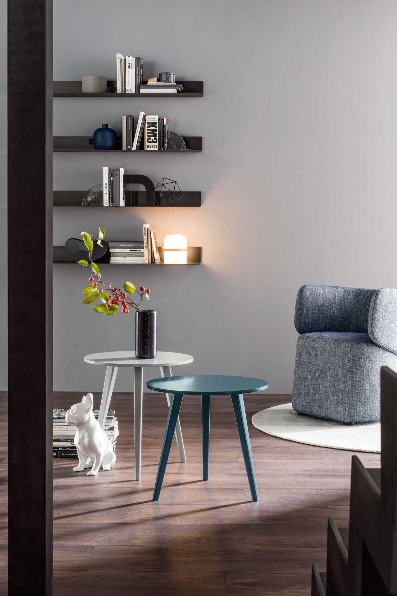 A stylish and modern living room features two round side tables in the foreground. The taller table is light gray and holds a dark, marble-patterned vase with a branch of red berries. The shorter table is a muted teal. To the left, a white ceramic dog figurine sits on a stack of books. In the background, a light blue-gray armchair is partially visible on a round rug. The wall behind the tables is a light gray, adorned with three long, black floating shelves. These shelves hold various books, decorative objects, and a small, glowing white lamp that casts a warm light. The room has dark wood flooring, and a dark vertical beam is visible on the far left.
