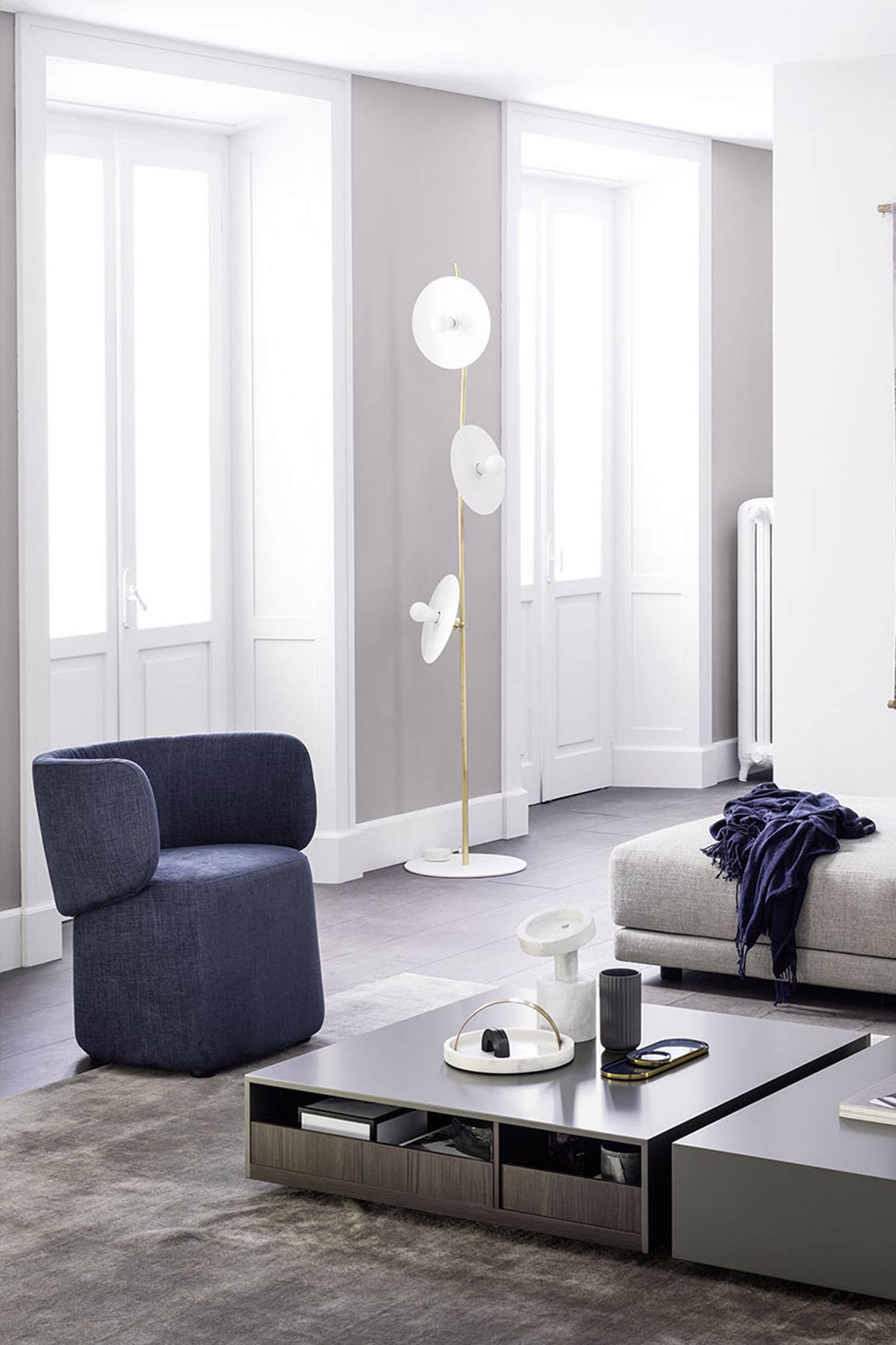 A modern living room features a dark blue upholstered armchair in the foreground, facing towards a multi-tiered, square coffee table with a dark gray top and drawers. On the coffee table, decorative items including a white tray, a white sculptural piece, and a black vase are arranged. To the right, a light gray ottoman or bench is visible with a dark blue throw blanket draped over it. In the background, against a light gray wall, a unique floor lamp with a gold stem and three circular white lights stands tall. Two large, white French doors or windows are on the back wall. The room is carpeted with a dark gray rug, and the floors around the rug are a dark wood.