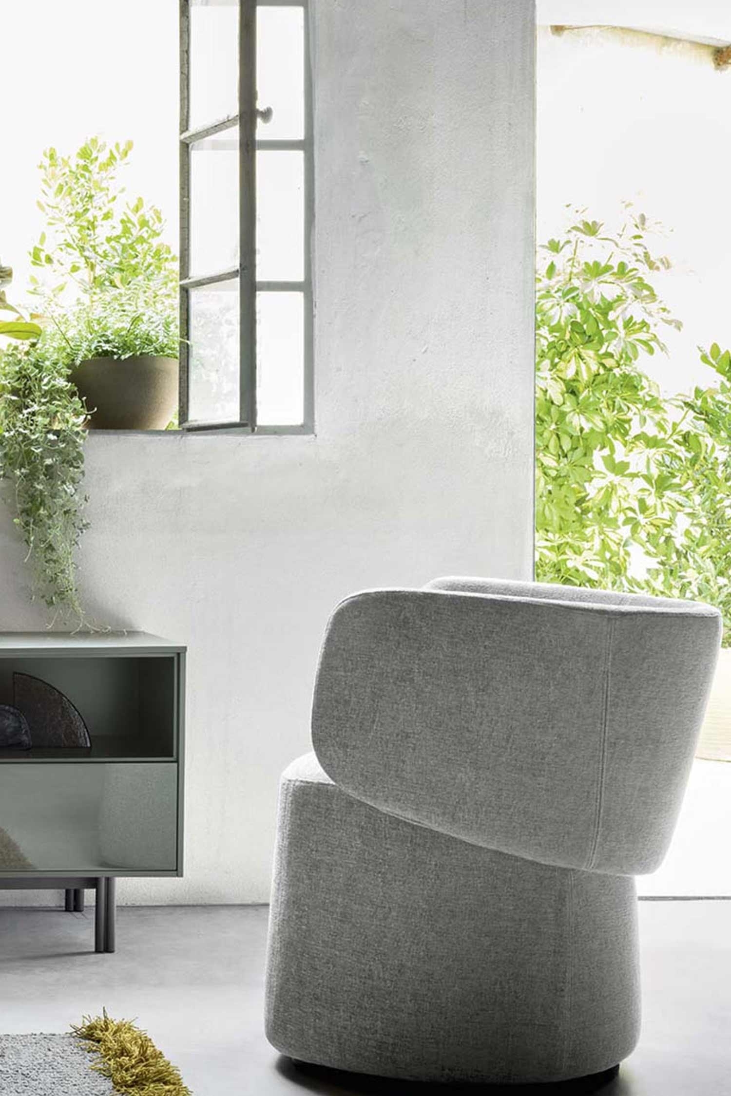 A full-height, professional studio shot of a light gray armchair with a distinct, minimalist design. The chair appears to be made of a textured, light gray fabric and has a two-part construction with a plush, rounded base and a curved backrest. The chair is positioned on a dark floor in a setting that resembles a rustic, sunlit room with a white, slightly textured wall. A large window with a dark metal frame is visible on the top left, and green plants are seen both on the windowsill and in the bright, outdoor space beyond the window. A dark gray modern console table is to the left of the chair.