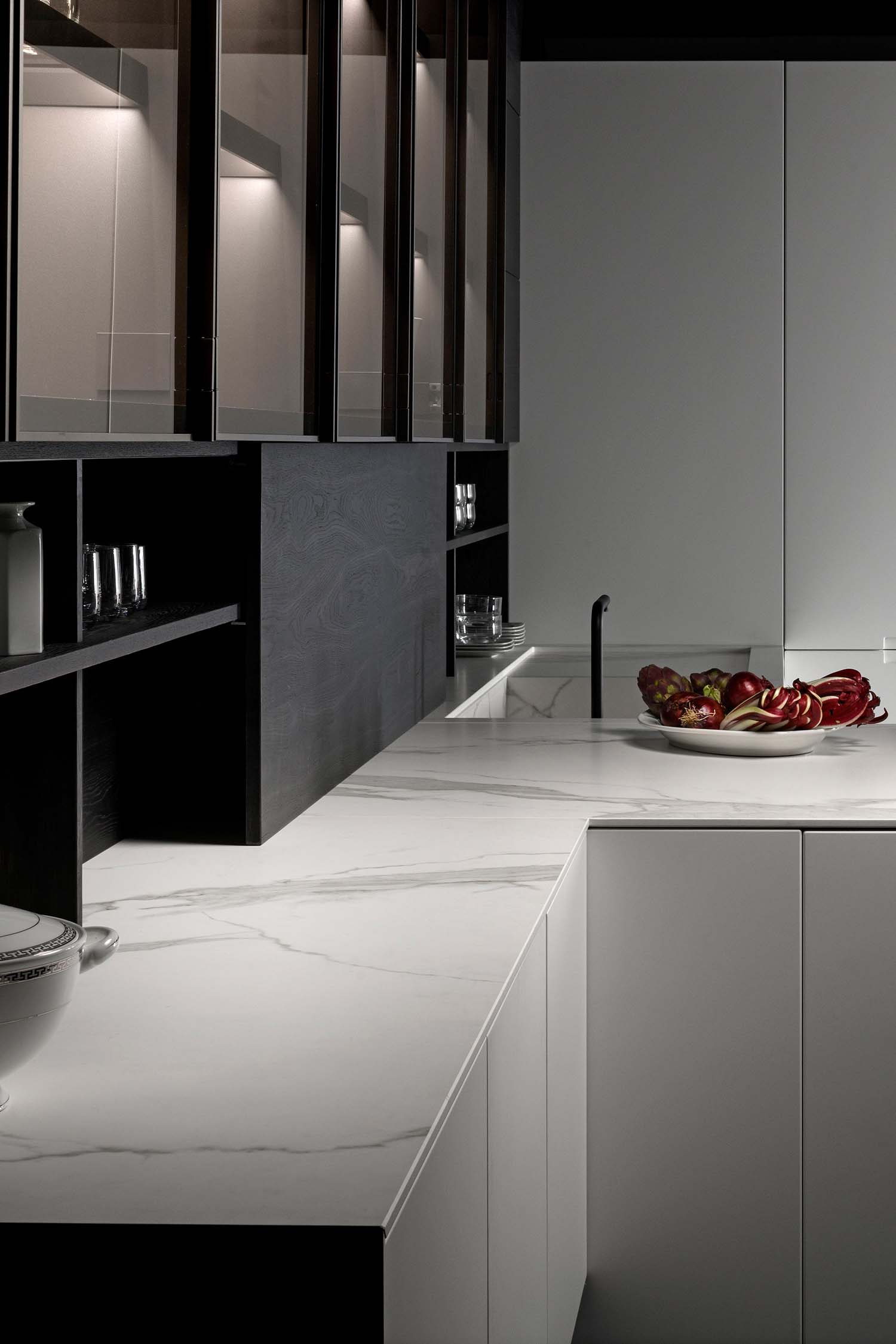 A detailed shot of a kitchen countertop with a marble-like pattern. The countertop is integrated with a sink and a black, modern faucet. The surface is clean and minimalist, with a bowl of fruit and an induction cooktop visible.