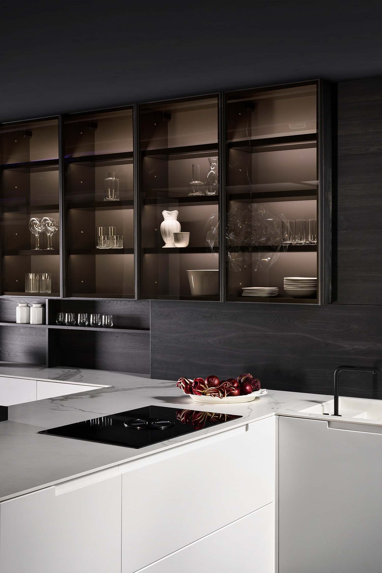 A close-up of a dark wood wall unit with smoked glass doors. The doors are hinged, revealing illuminated shelves displaying various dishes, glasses, and decorative items. The contrast between the dark wood and the backlit interior creates a sense of luxury.