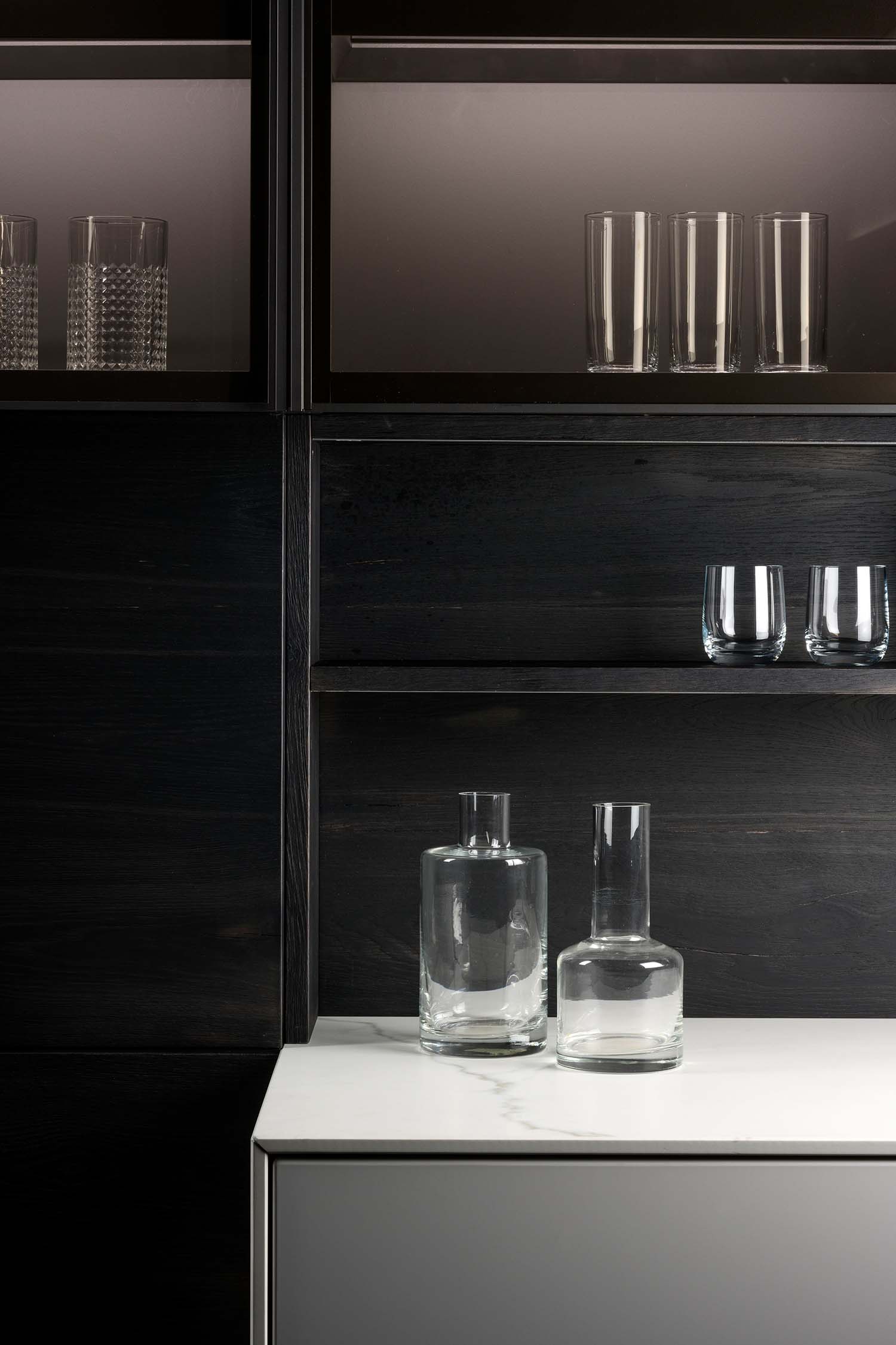 A shot of the open shelving unit within the dark wood kitchen cabinetry. The image shows several empty glass tumblers on a lower shelf and two clear glass vases on the white countertop below. The texture of the wood is a central feature.