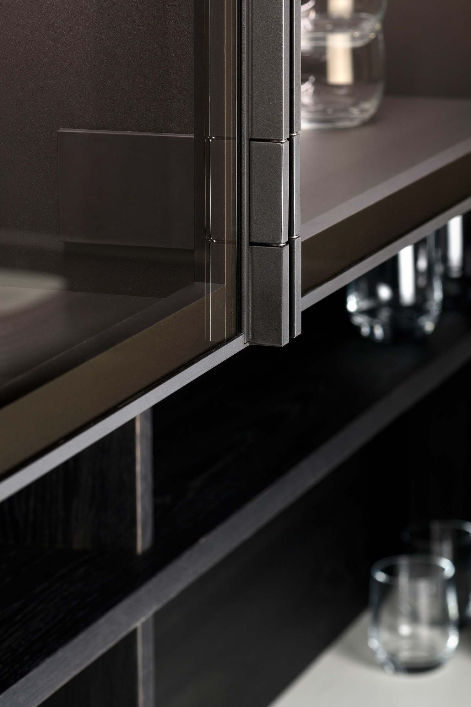 An extreme close-up of a cabinet hinge on a glass-fronted display door. The hinge has a contemporary matte dark grey or black finish, emphasizing the quality and attention to detail in the kitchen's construction.
