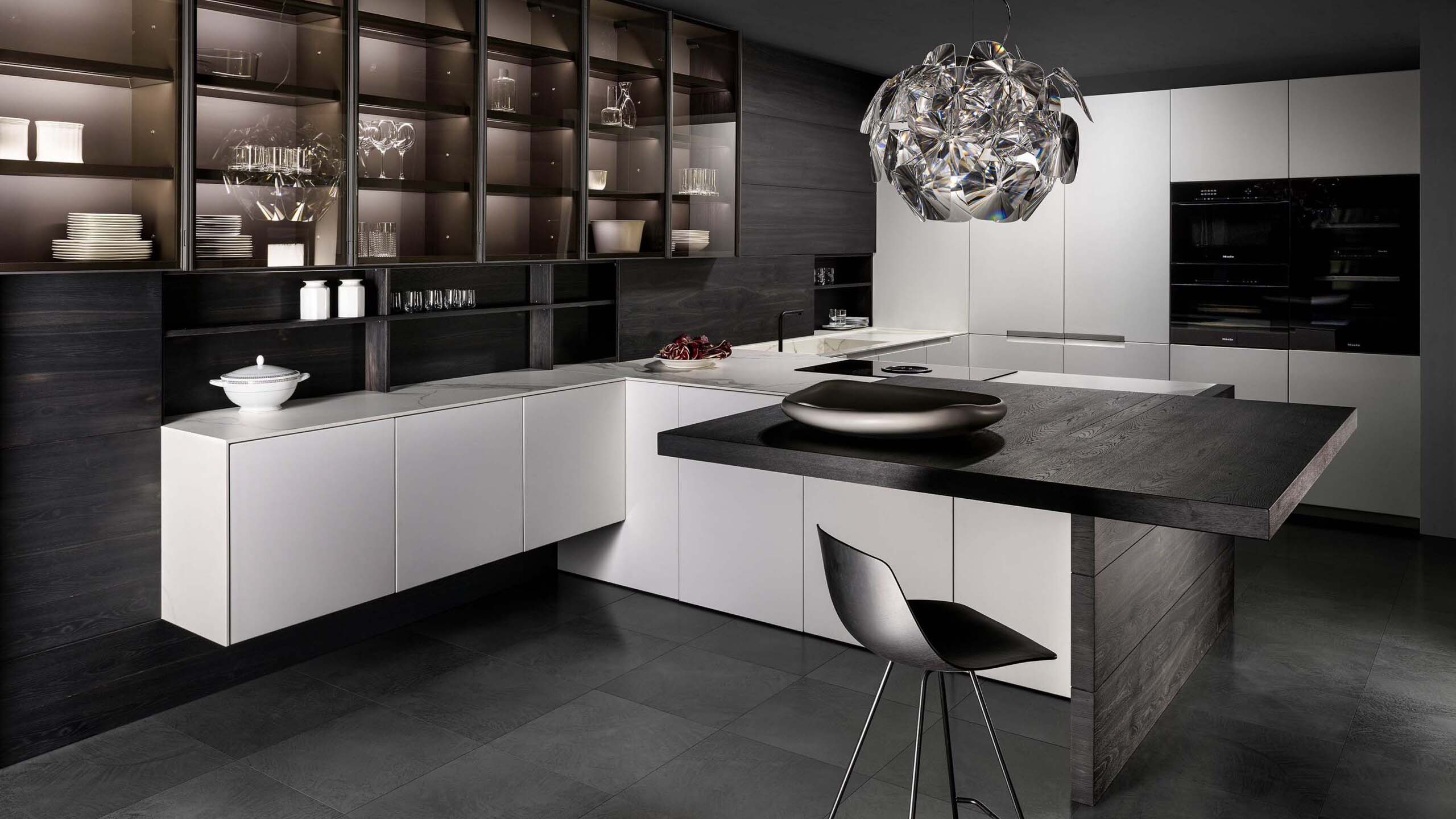 A wide shot of a luxury, handleless kitchen with a dramatic contrast between light and dark elements. The space features a large L-shaped island with a dark wood block that extends to a breakfast bar with a single black stool. The main kitchen cabinets are matte white, while a large wall unit with integrated lighting is made of dark wood and glass-fronted display cabinets.