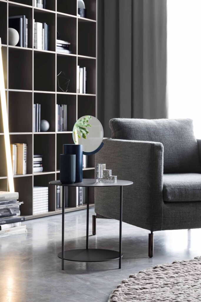 A cropped shot of a gray upholstered armchair with a wooden base and black metal legs. In front of the chair is a two-tiered black metal side table holding a dark blue cylindrical vase, a few clear decorative pieces, and a small round mirror reflecting a green plant. Behind the table, a tall modular bookshelf filled with books and various objects stands against the wall. A tall, thin light fixture with a golden glow is visible on the far left. The room has a concrete floor with a portion of a round gray rug.