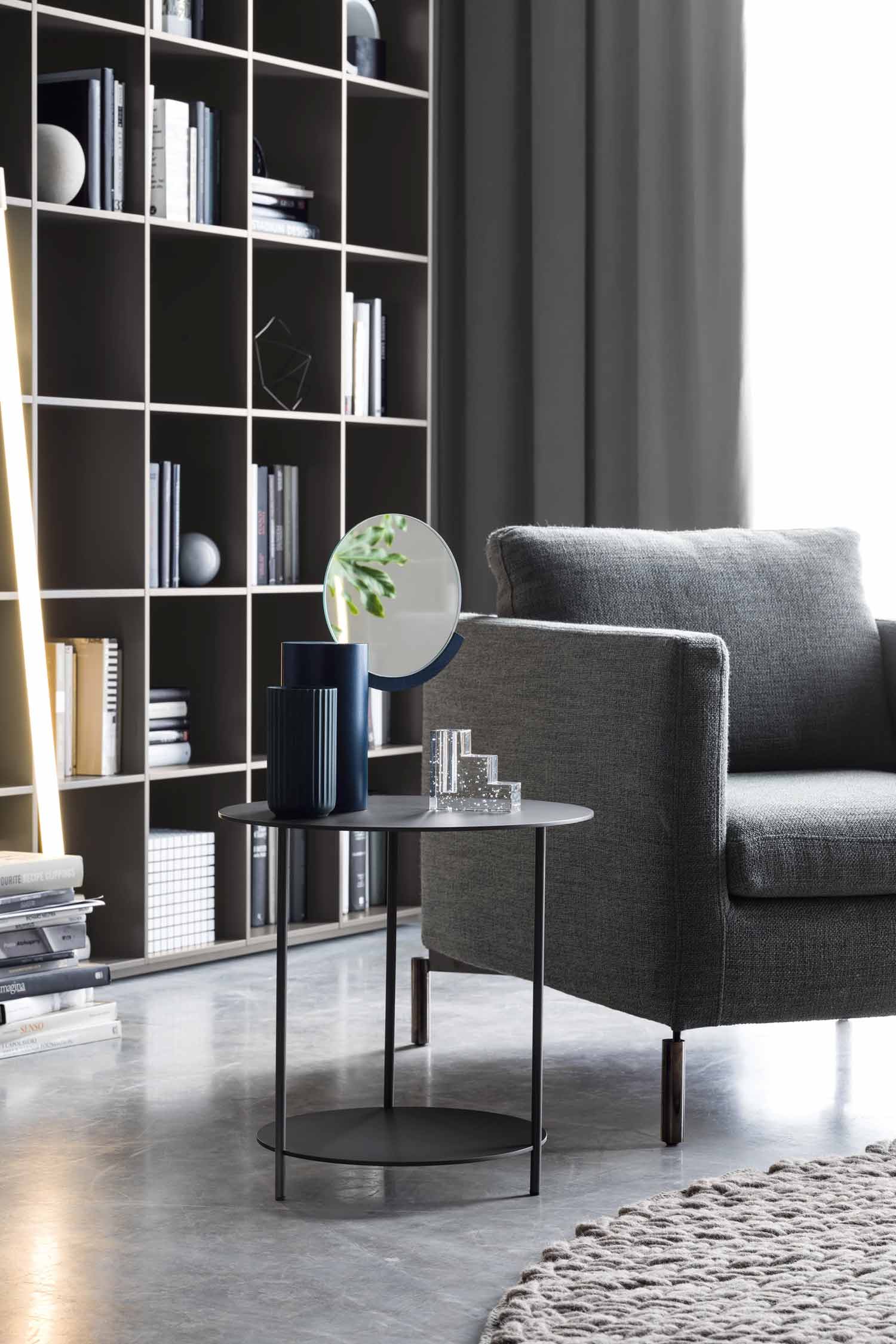 A cropped shot of a gray upholstered armchair with a wooden base and black metal legs. In front of the chair is a two-tiered black metal side table holding a dark blue cylindrical vase, a few clear decorative pieces, and a small round mirror reflecting a green plant. Behind the table, a tall modular bookshelf filled with books and various objects stands against the wall. A tall, thin light fixture with a golden glow is visible on the far left. The room has a concrete floor with a portion of a round gray rug.