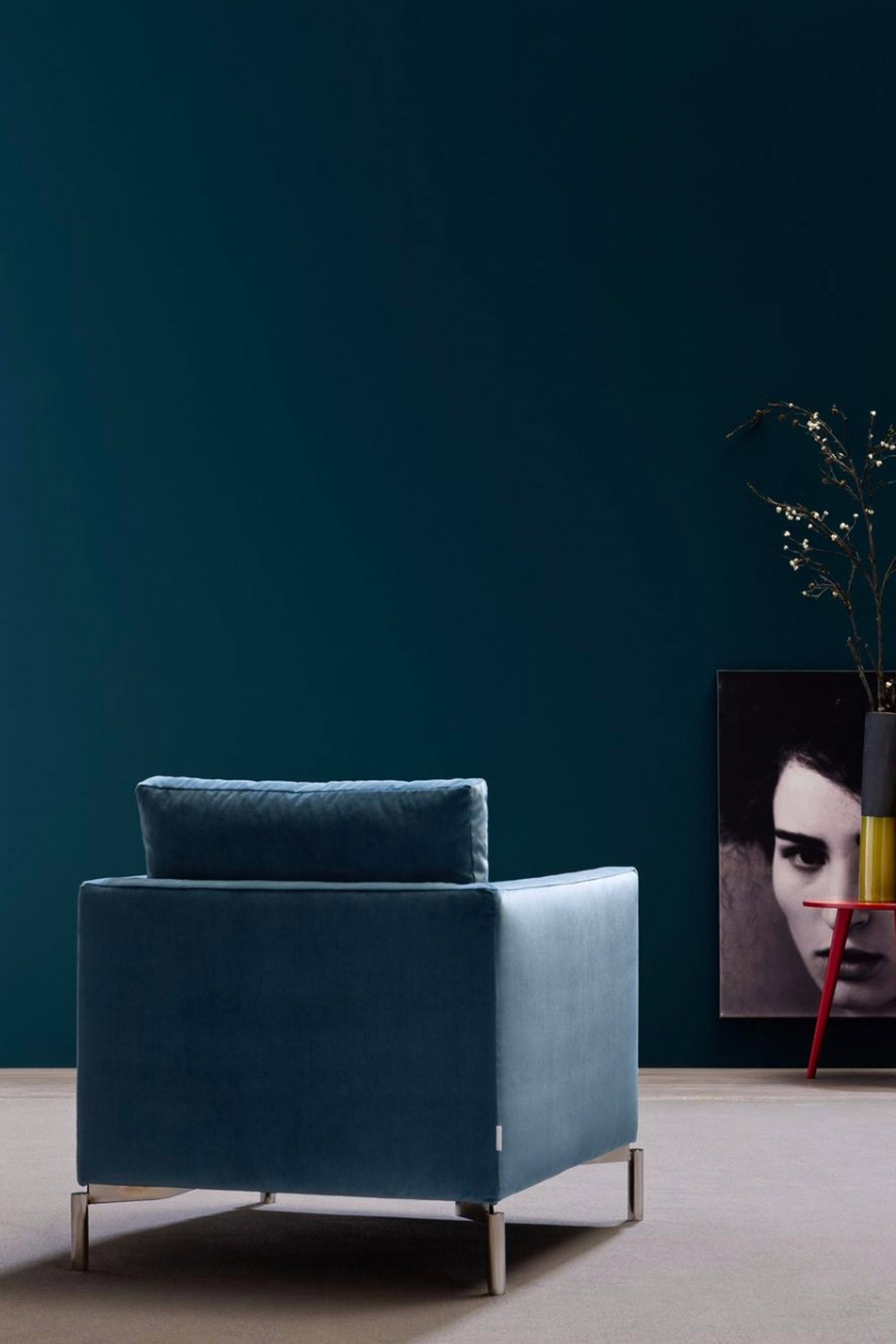 A rear-view of a modern armchair upholstered in dark blue velvet fabric, featuring a square silhouette and thin silver metal legs. The chair is placed against a solid teal-blue wall. To the right, a framed black and white portrait of a woman with red lips is propped against the wall. A small red and yellow side table next to the portrait holds a vase with bare branches.