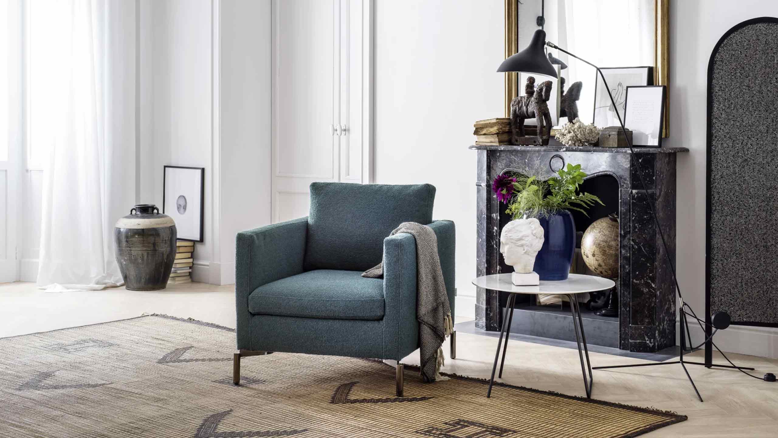 A blue upholstered armchair with thin bronze legs and a gray throw blanket is placed in a cozy, elegant living room. To the right, a round white marble side table holds a white sculptural bust, a large blue vase with flowers and greenery, and a globe. Behind the armchair is a black marble fireplace mantel adorned with books, a horse sculpture, and a black desk lamp. Above the mantel, a large mirror with a gold frame reflects the room. A large decorative vase sits on a stack of books to the left of the chair. The room has light wood flooring with two overlapping rugs, and a tall, arched black screen stands on the far right.