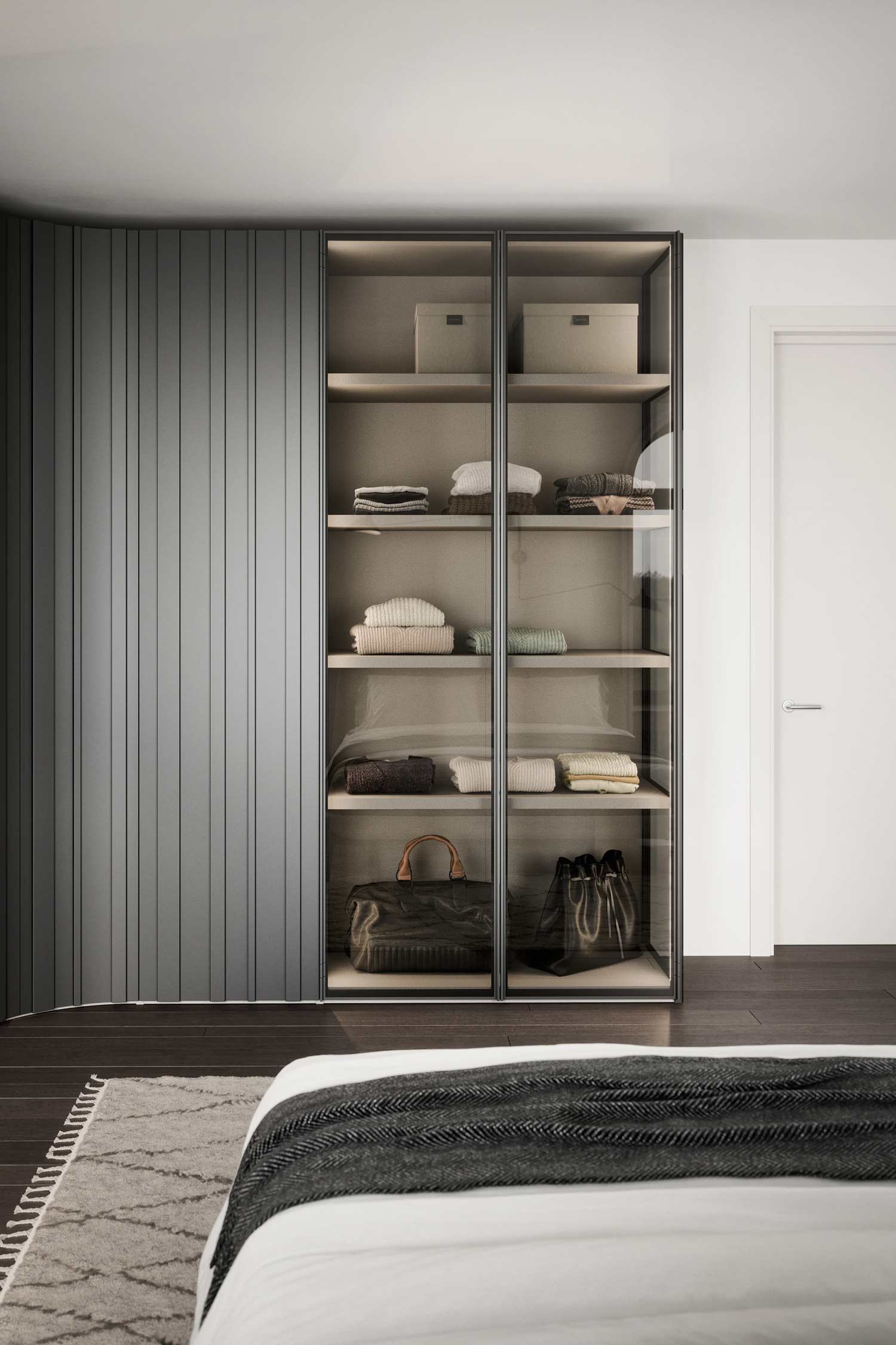 A side view of a modern wardrobe system in a bedroom. The wardrobe features a curved section with gray, slatted doors next to a glass-fronted section with illuminated shelves displaying folded clothes and bags.