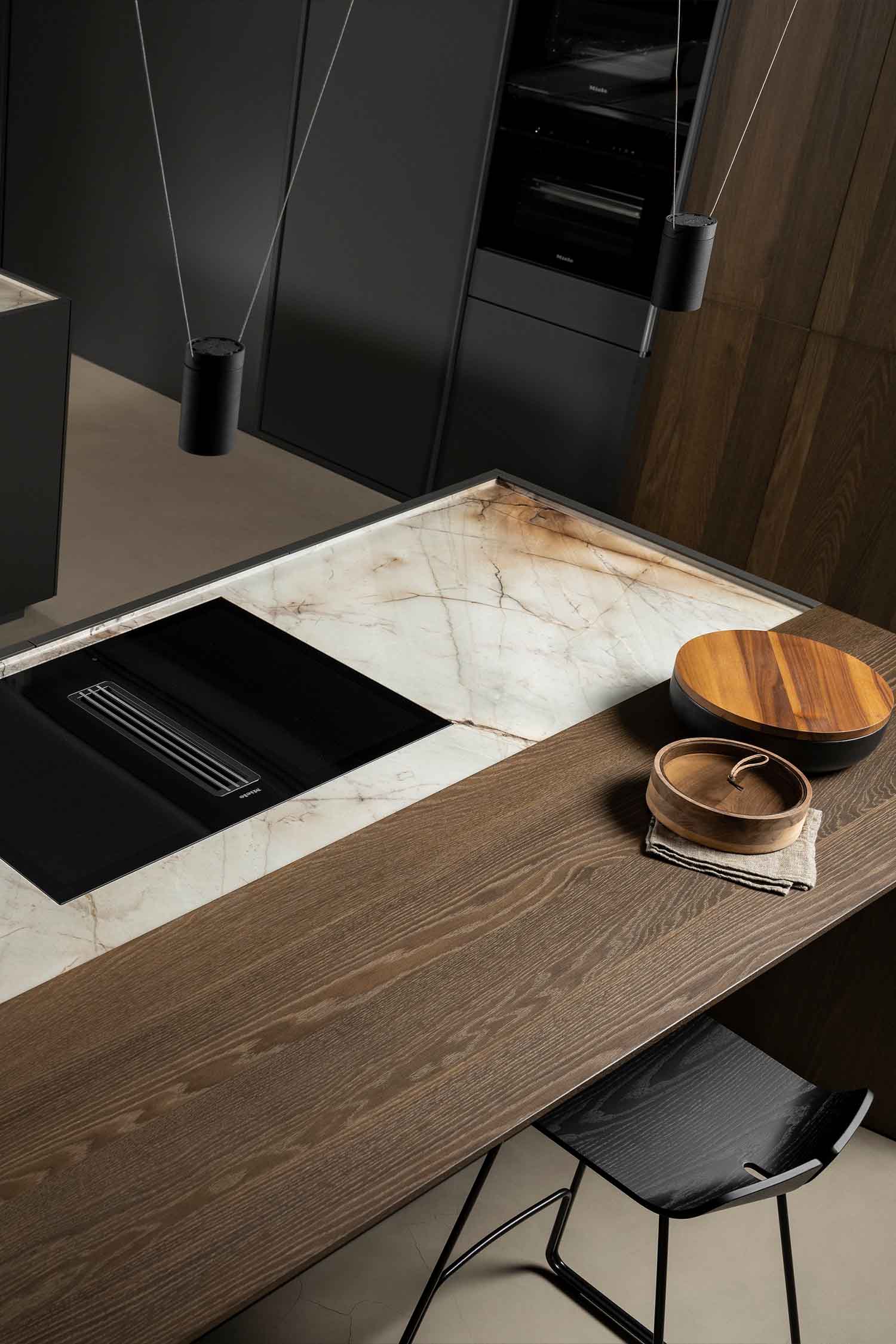 An overhead, angled shot of a modern kitchen island. One side features a black induction hob with an integrated extractor, set into a striking white marble countertop. The other side is a waterfall-style breakfast bar made of dark wood veneer. A black stool is tucked underneath.