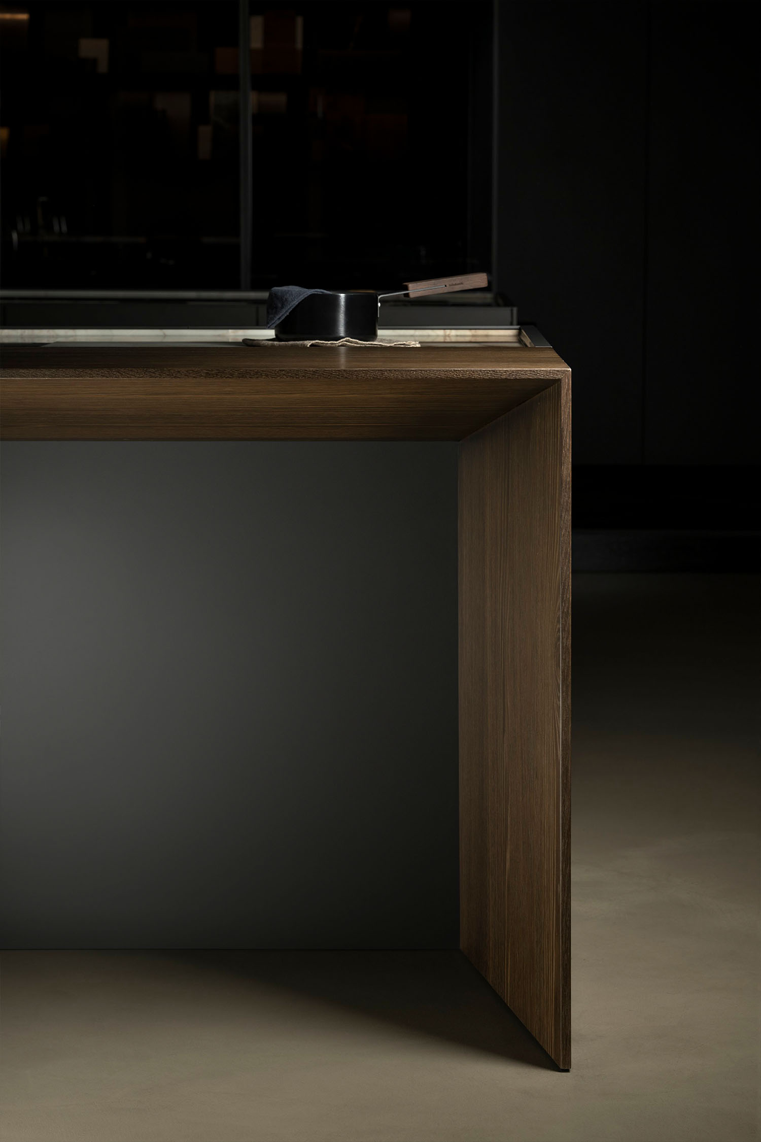 A low-angle shot focusing on the end of a kitchen island. The island features a waterfall-style top and side panel made of dark wood veneer, contrasting with a smooth, grey side panel.