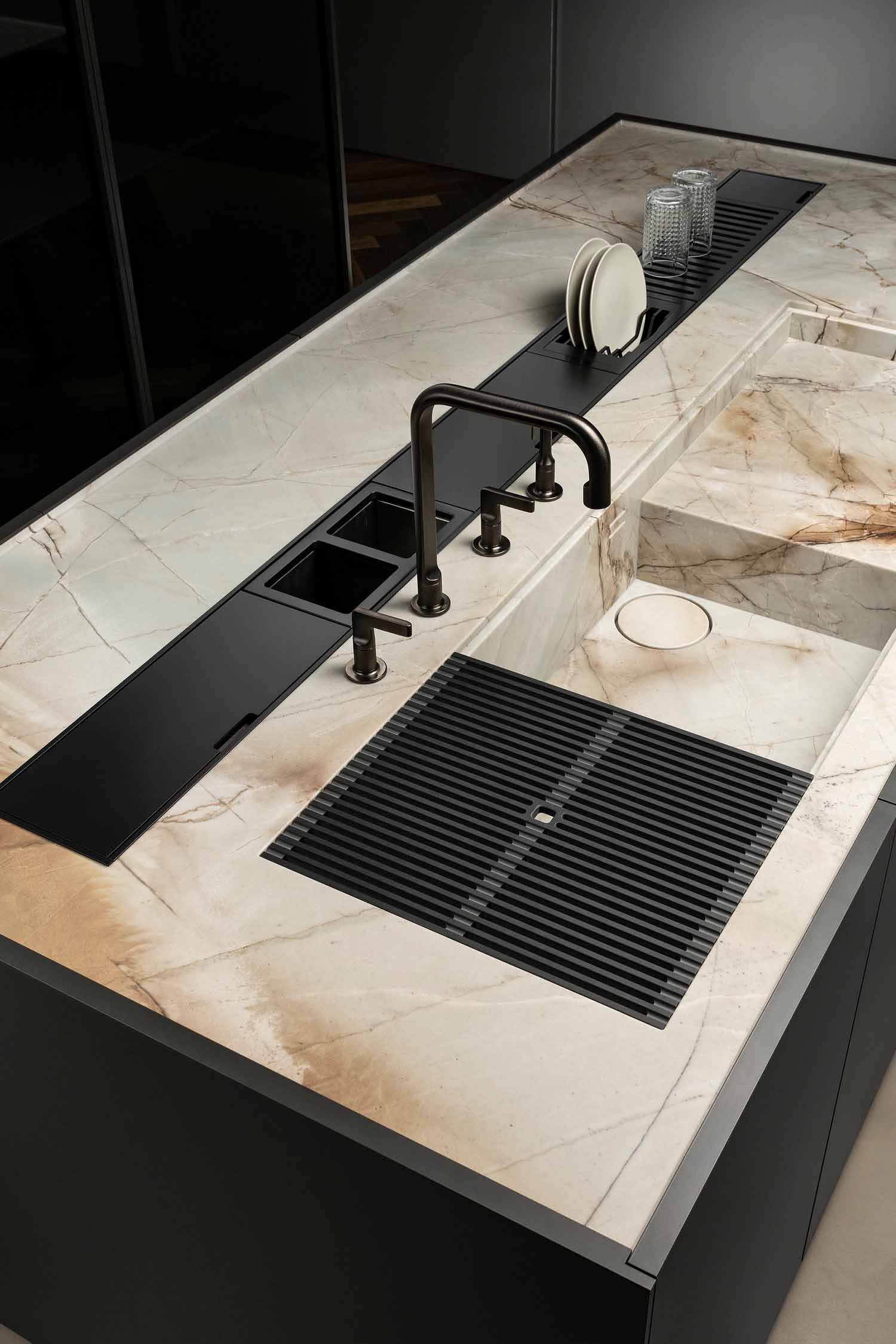 A close-up of a kitchen island countertop made of light-colored marble with prominent dark veining. It features a modern, integrated black sink with a sleek black faucet and a removable, grooved drying rack. A black cover conceals a section to the left of the sink.