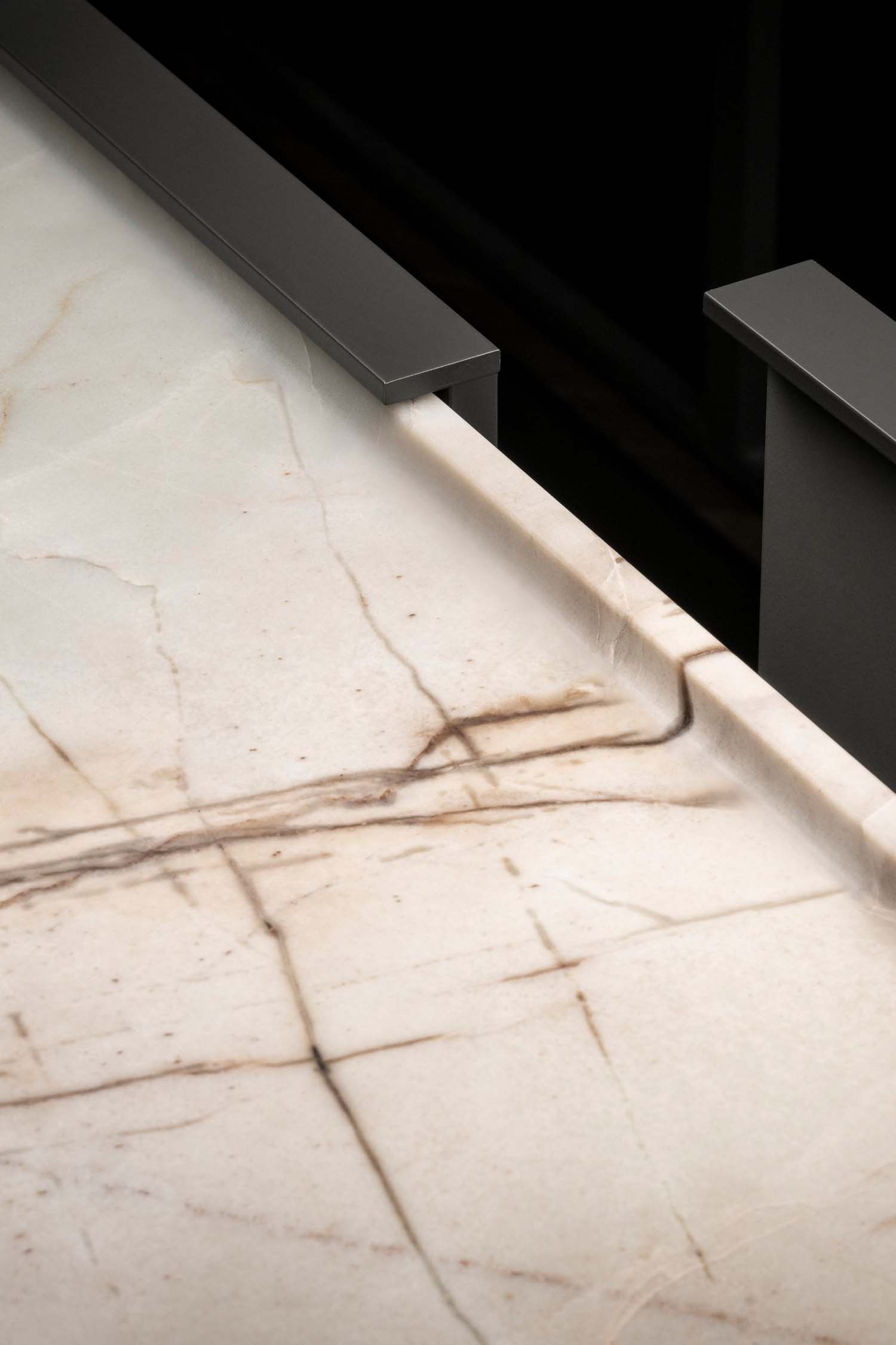 A very close-up detail of a light-colored marble kitchen countertop with brown and grey veining. The edge of the countertop meets a dark metal frame.