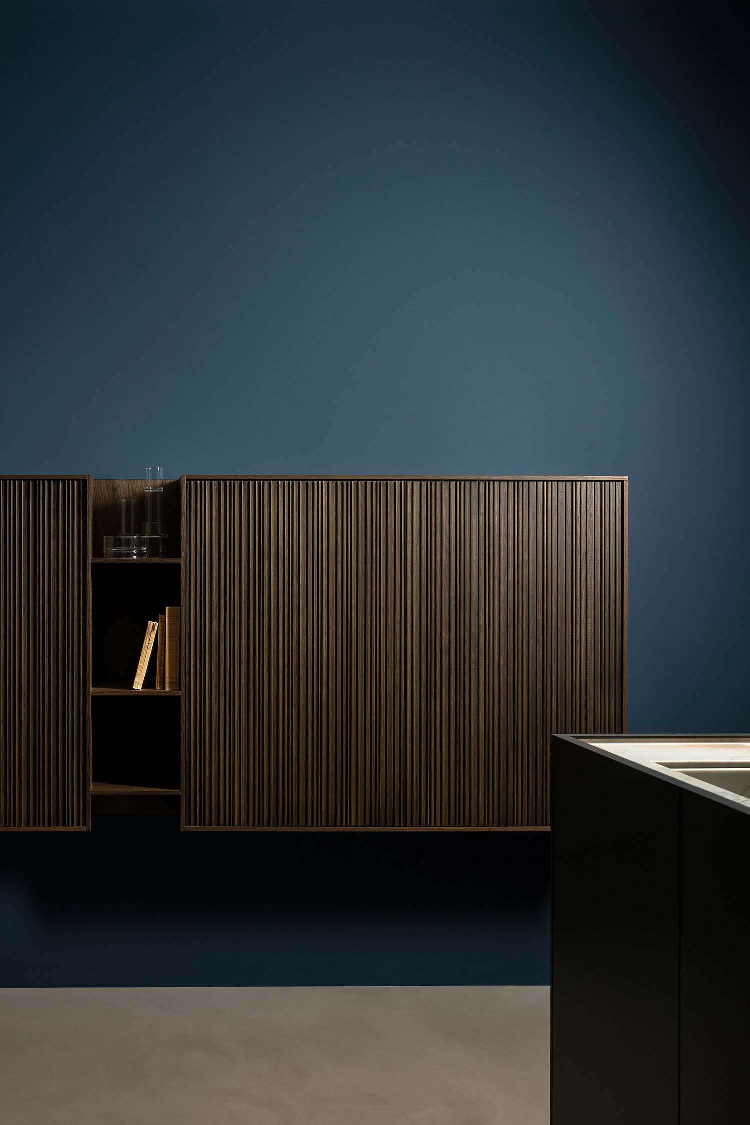 A detailed shot of a wall-mounted wooden cabinet against a deep blue wall. The cabinet has a ridged, slatted texture and appears to have a single open shelf revealing a book and a glass vase.