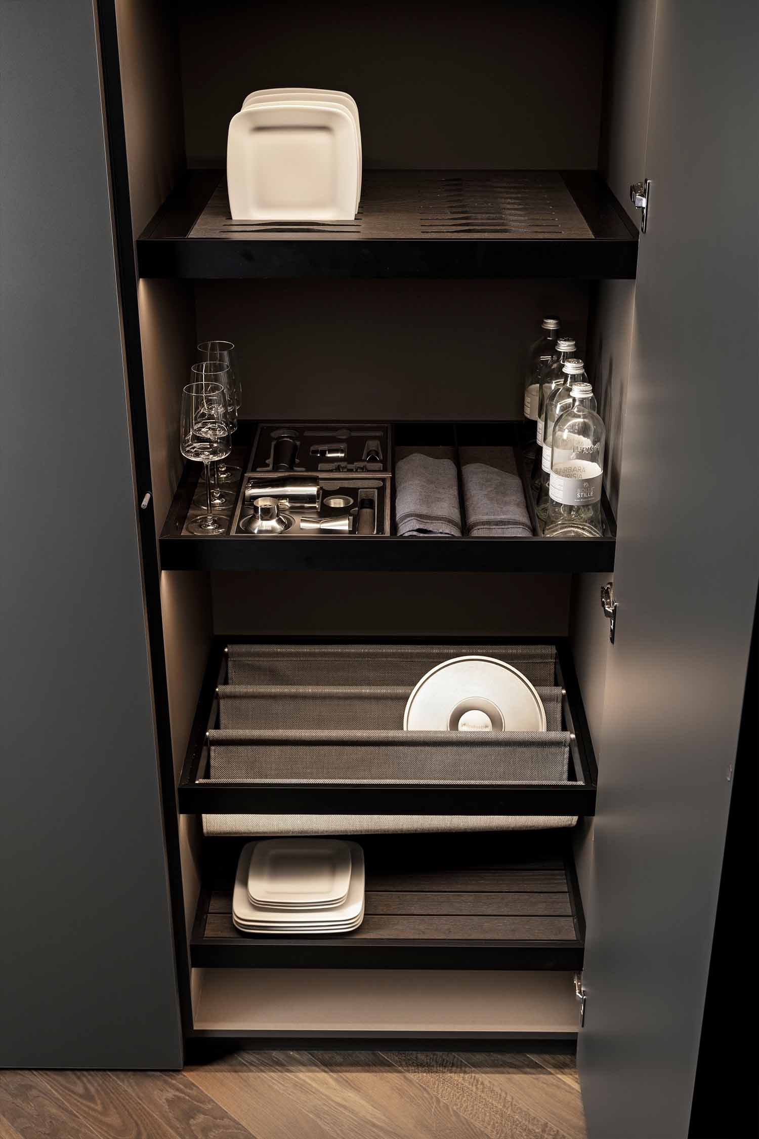 A close-up view of an open luxury kitchen pantry cabinet with integrated lighting. The shelves hold organized kitchenware, including a stack of white plates, glassware, a cutlery tray, and folded towels. A pull-out storage drawer below holds a white bowl.