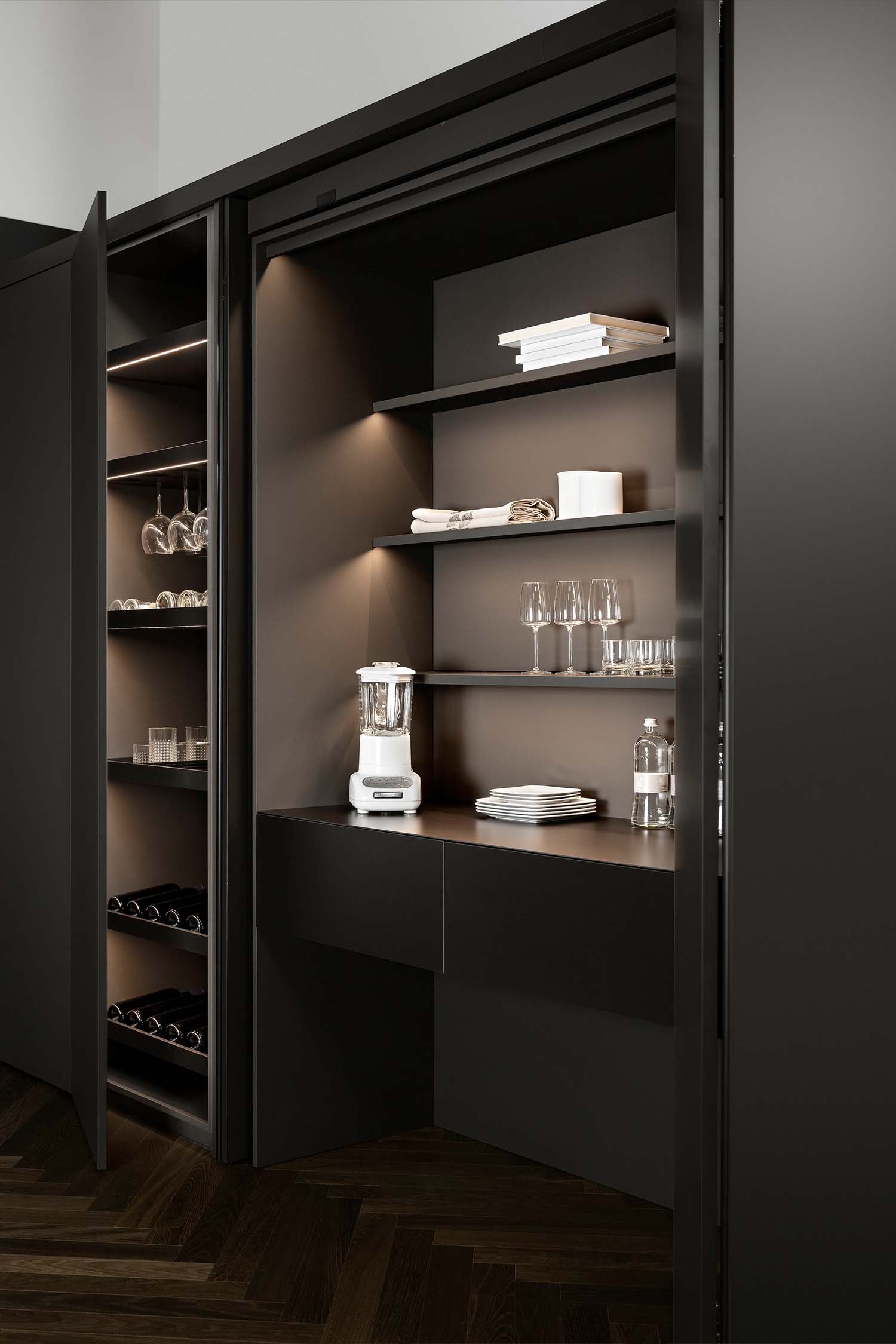 A detail of a dark kitchen pantry with open doors. The interior is a dark matte finish with subtle, warm integrated LED lighting. Shelving holds a blender, glasses, a stack of white plates, and other kitchen items. A wine rack is visible on the far left.