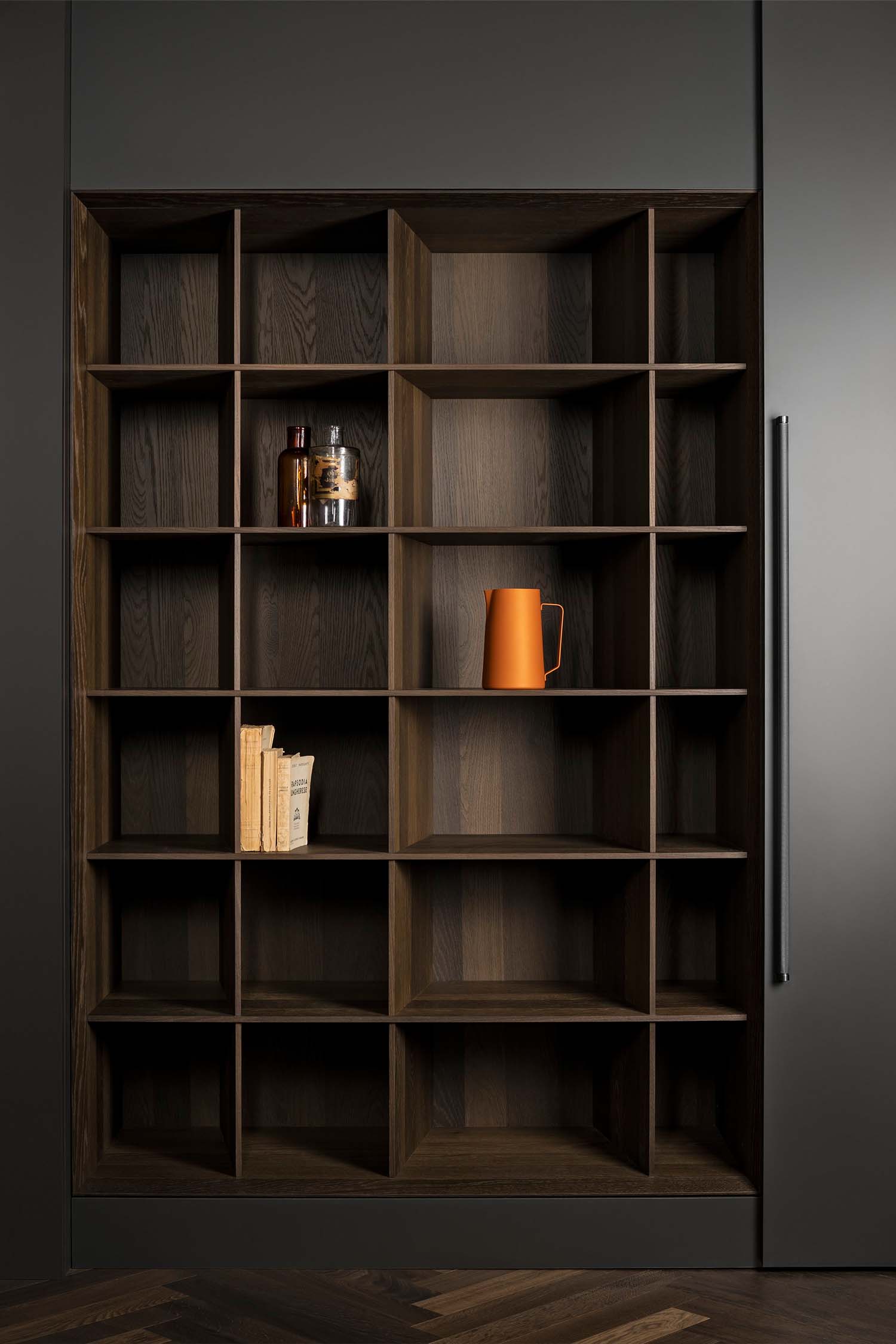 A large, dark wood veneer shelving unit with a grid-like pattern of square and rectangular compartments. The shelves are mostly empty, with a few decorative items like books and an orange pitcher.