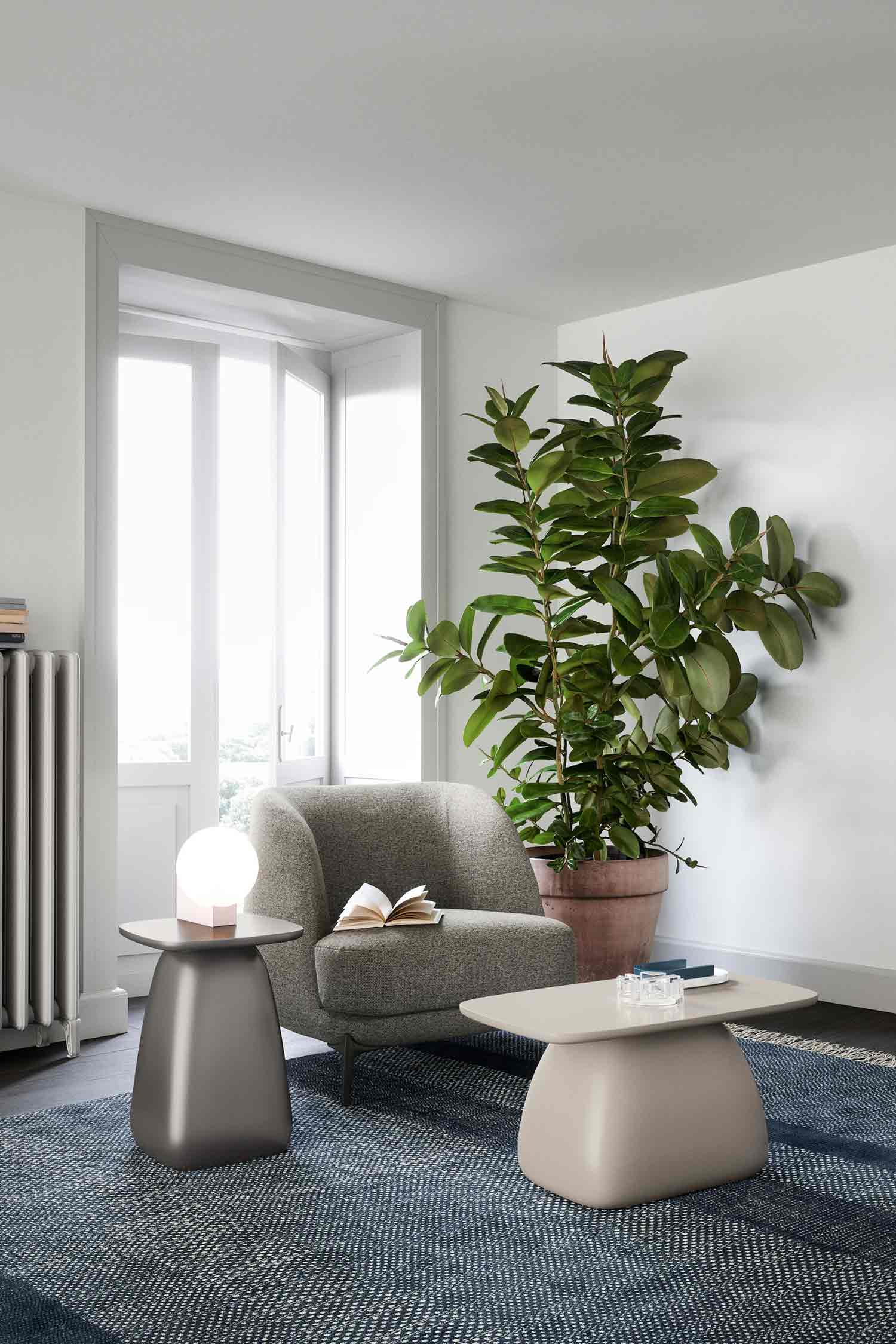 A gray upholstered armchair sits on a blue and white woven rug in a brightly lit room. An open book rests on the seat of the chair. A large potted plant with broad green leaves stands behind the armchair. To the left, a tall, bronze-colored side table with a unique curved shape holds a glowing pink lamp. A second, light-colored side table sits in front of the chair, holding a stack of books and a small clear box. A tall white window and a radiator are visible on the left side of the room.