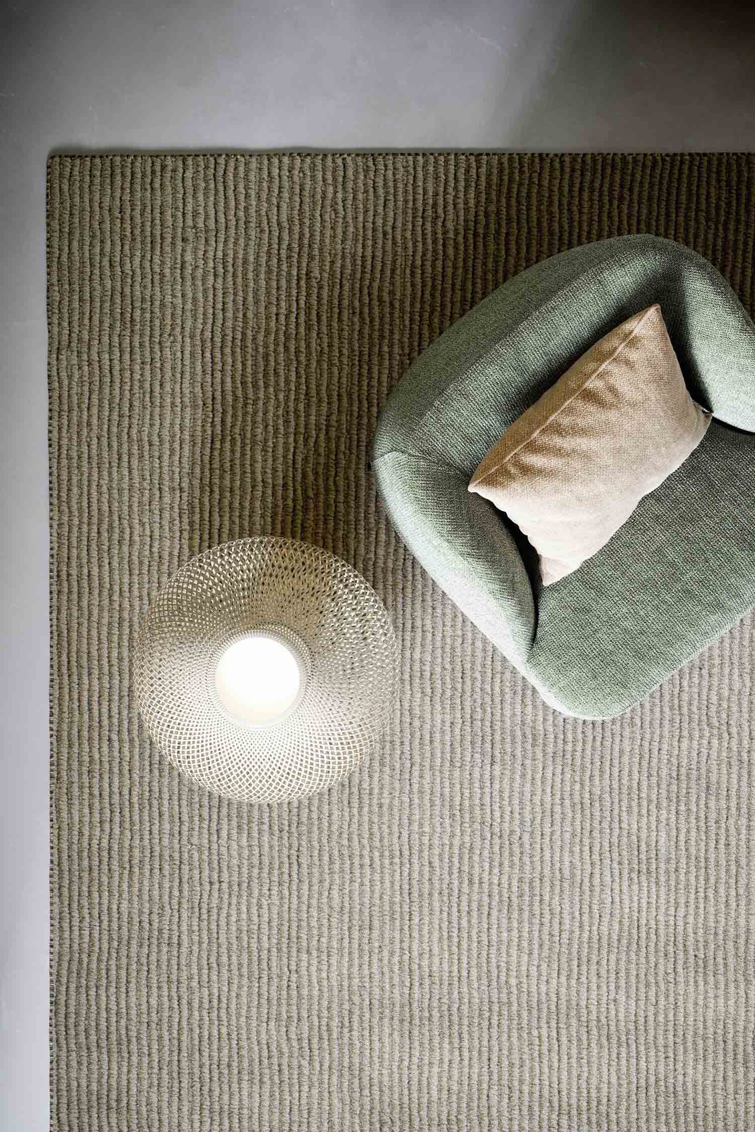A top-down view of a light green textured armchair with a small, light-colored rectangular pillow on it. To the left, a modern, light-colored lamp with a unique woven, beehive-like shade is glowing. Both the chair and the lamp are on a light beige, ribbed rug.