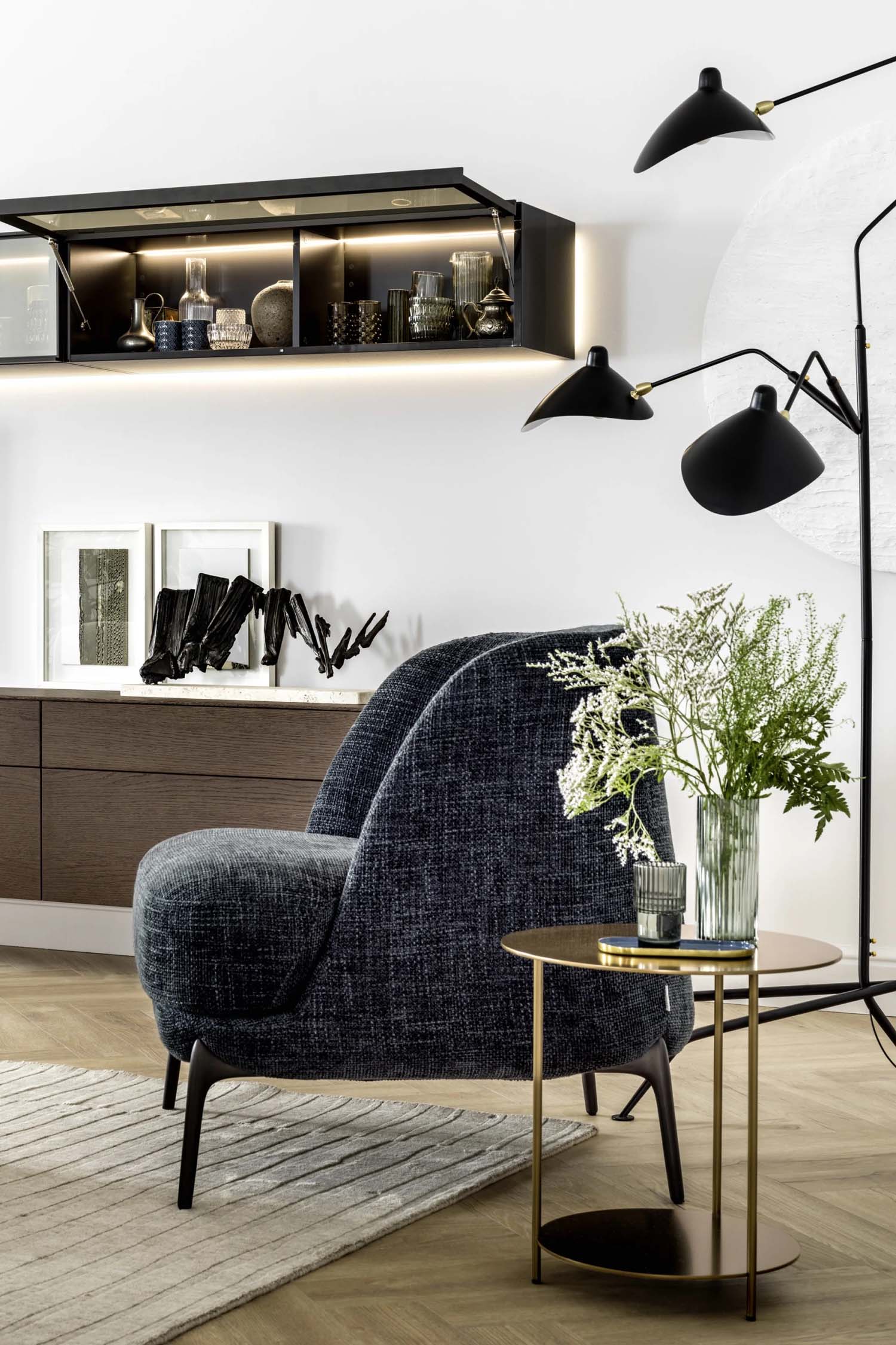 A close-up shot of a luxurious, contemporary armchair. The armchair is upholstered in a soft, light-colored fabric and features a unique curved armrest with a distinctive seam detail. The photo highlights the texture of the fabric and the fine craftsmanship of the Italian design.