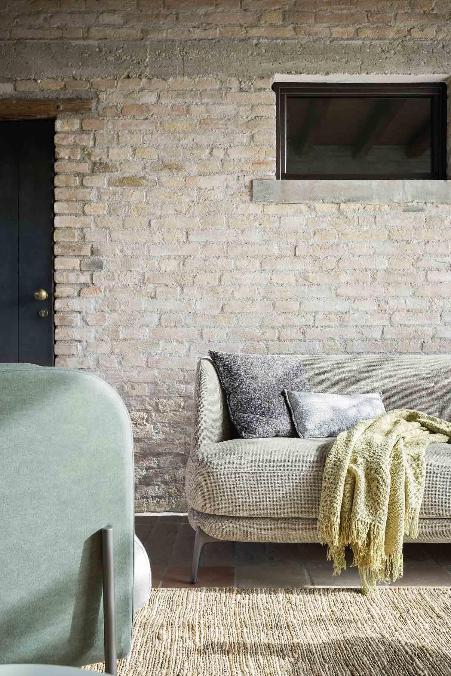 A close-up shot of a beige-colored sofa with two gray cushions and a light green blanket. The sofa sits in front of an aged brick wall next to a light-colored armchair.