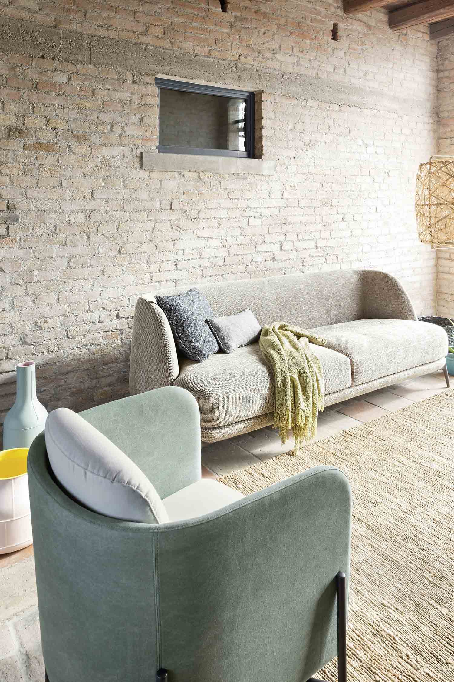 A full shot of a beige sofa with a light green blanket and two gray pillows. The sofa is in a room with a light brick wall, a prominent light green armchair, and a woven rug.