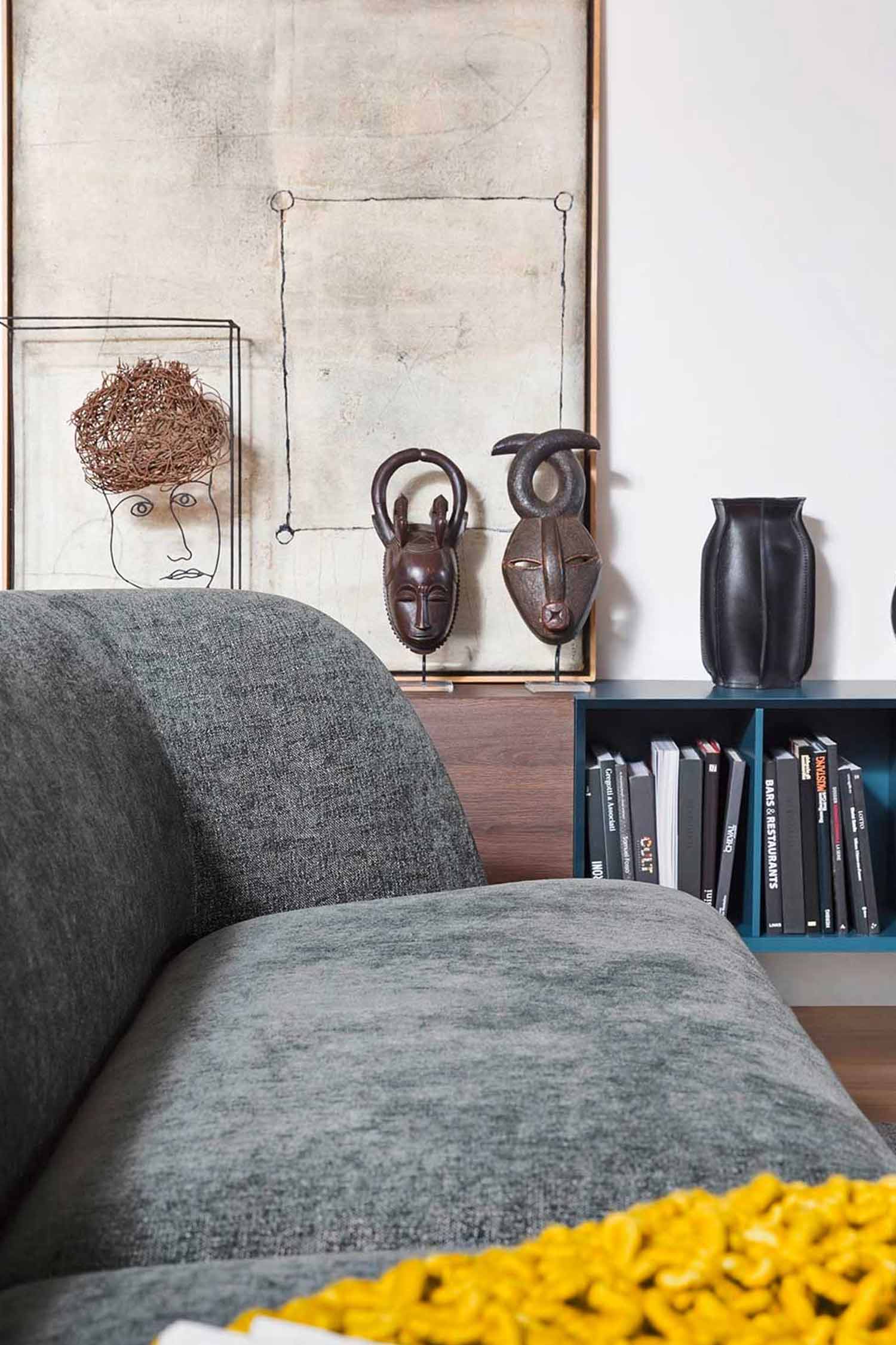 A side view of a dark gray sofa with a bright yellow blanket draped over it. In the background, there's a blue bookshelf, a wooden cabinet with two carved African masks, and modern art pieces on a wall.
