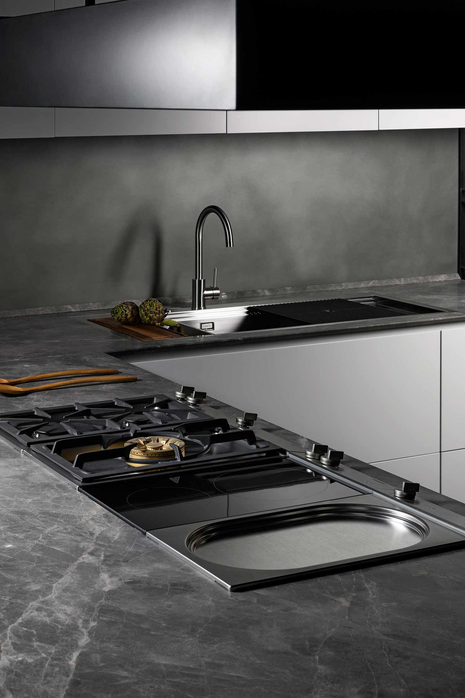 A close-up of a kitchen countertop and sink area. The countertop is a dark grey, veined marble, and it features a flush-mounted stainless steel sink with a modern chrome faucet. To the left is a gas cooktop with multiple burners, and to the right is a small, built-in metal basin.