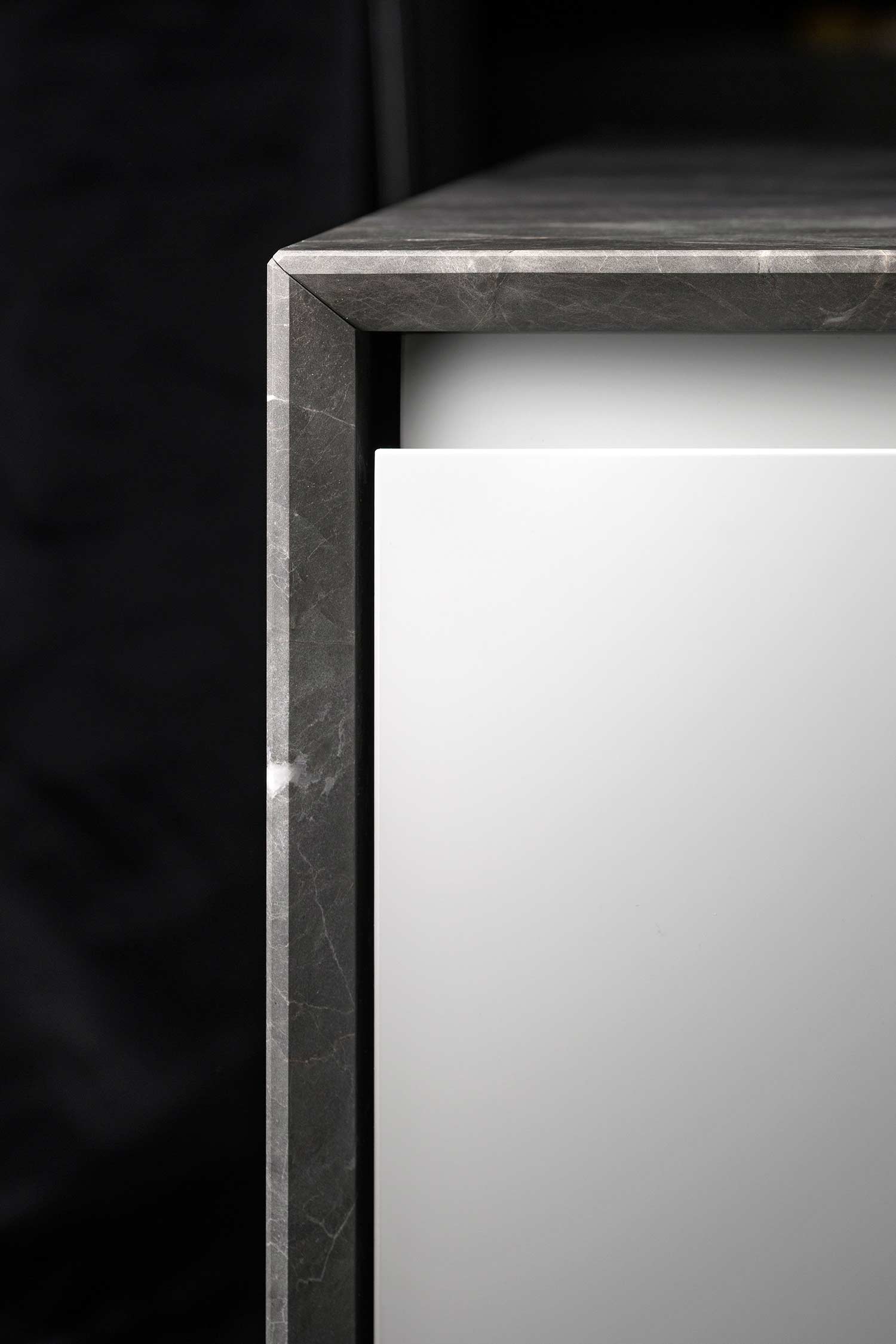 A detailed shot focusing on the corner of a kitchen cabinet. The image highlights the seamless joint of the dark grey, veined marble countertop, showing how the material wraps around the edge to create a thick, clean line. The marble frames a sleek white, handleless cabinet door.