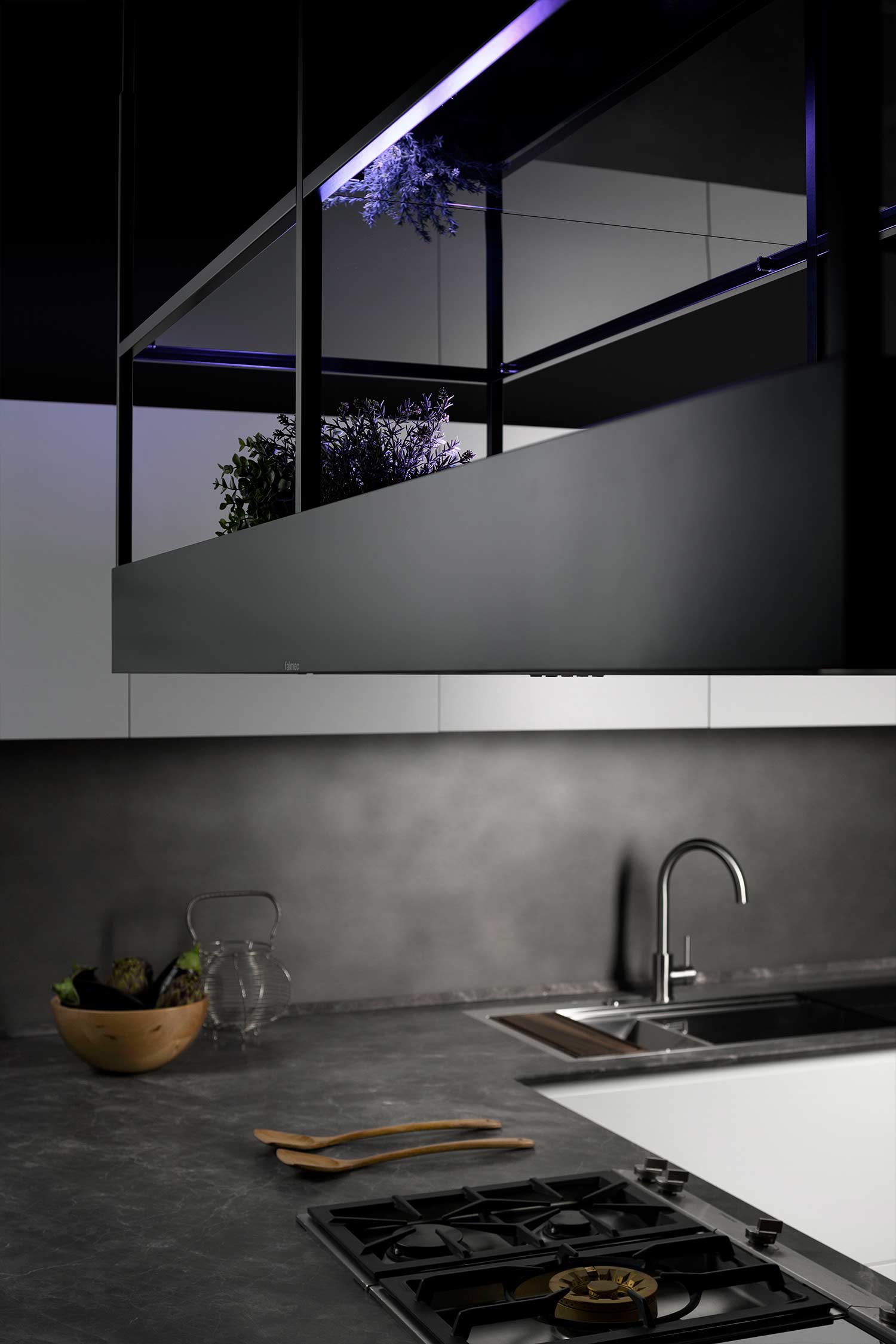 A detailed shot of the unique, suspended black metal structure above the kitchen island. The structure serves as both a range hood and a lighting feature, with integrated purple and white lights illuminating small potted plants placed on a shelf within the frame. A simple metal faucet is visible on the counter below.