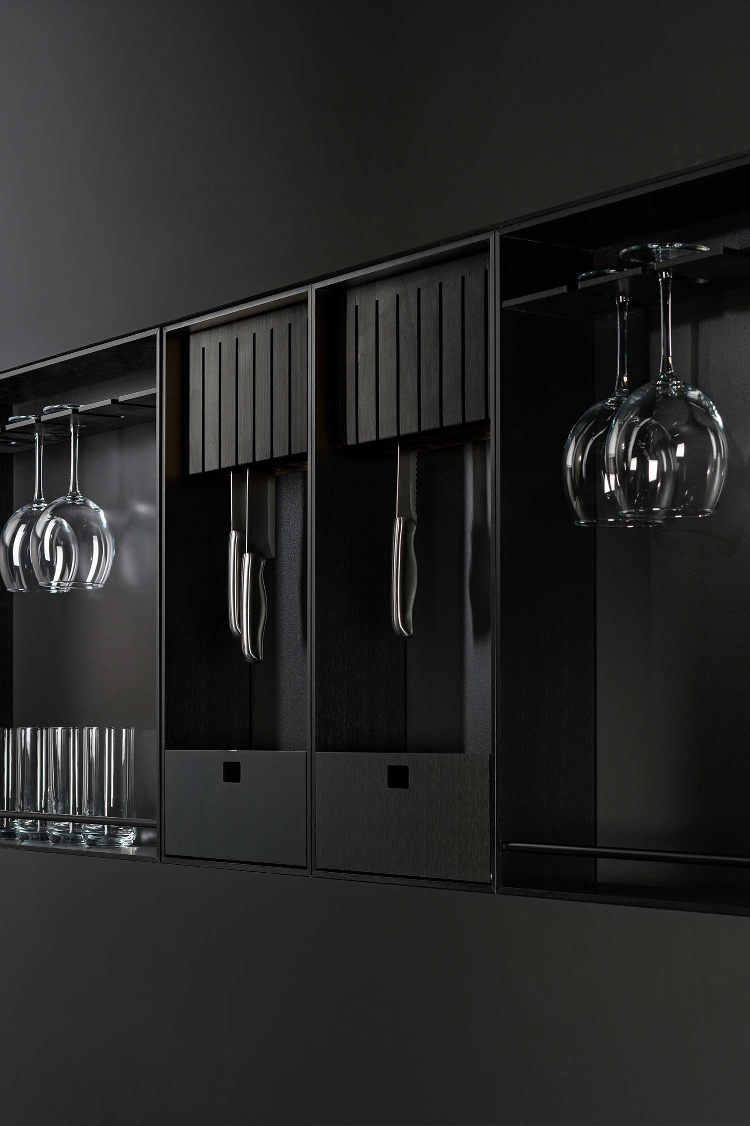 A close-up of a built-in black wall unit in a contemporary kitchen. The unit features dedicated compartments for kitchen tools, including a suspended knife block and built-in racks for wine glasses and tumblers.