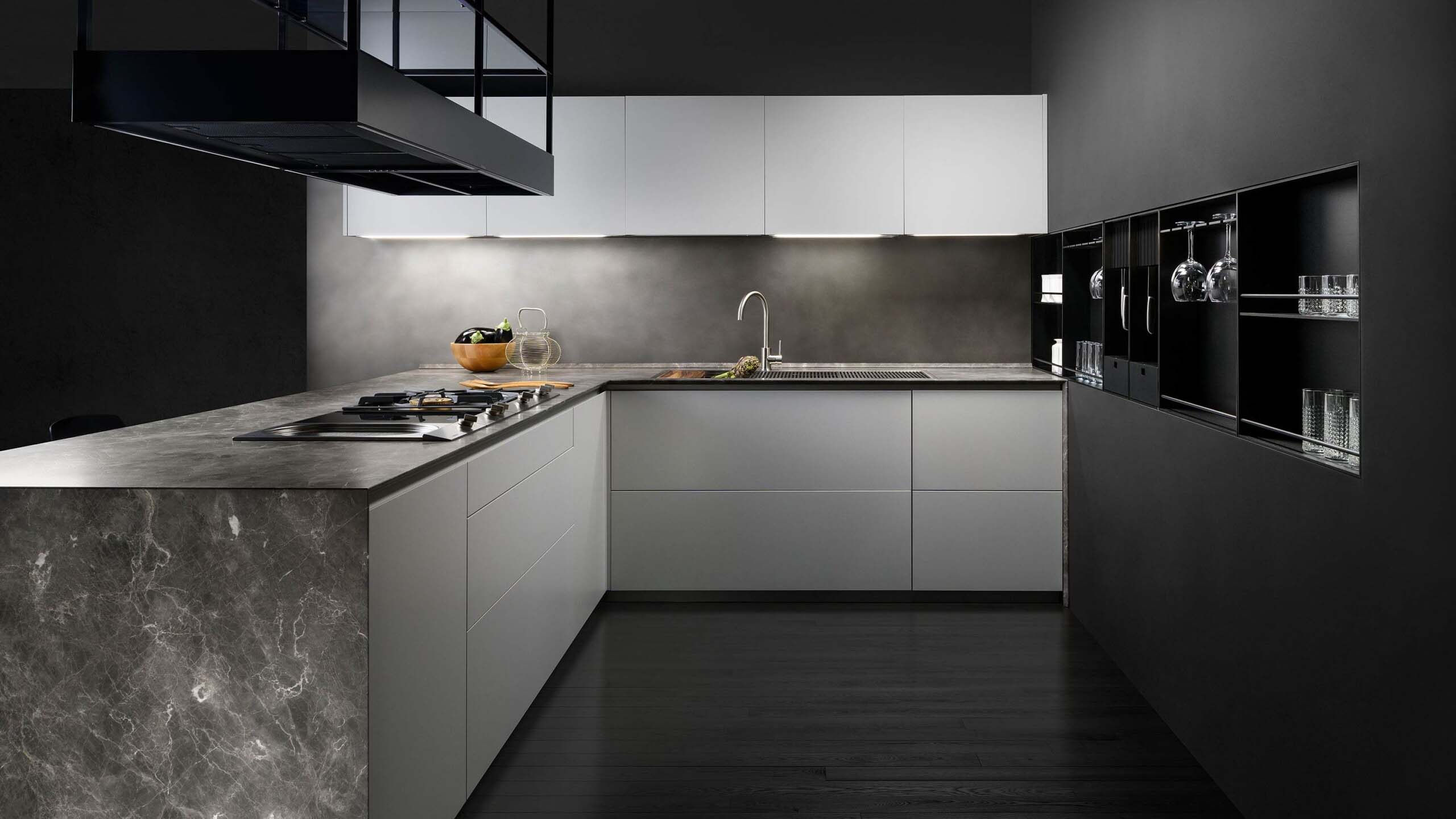 An overview shot of a contemporary kitchen island with seating. The kitchen island has a dark grey marble countertop that extends into a wooden-edged breakfast bar. Two black bar stools are tucked underneath. The island incorporates a cooktop and a circular prep area. A black metal structure with integrated plants and lighting hangs above, and a built-in wall unit showcases glassware and a knife block.