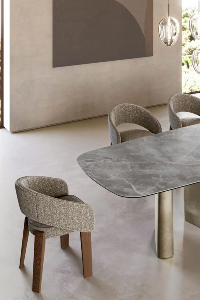 A close-up, high-angle view of a modern dining table's corner and a matching chair. The table has a dark grey marble top with subtle reddish-brown veining and a champagne-colored cylindrical metal base. The chair is upholstered in a textured, light grey-brown fabric with a curved back and sits on dark wooden legs.
