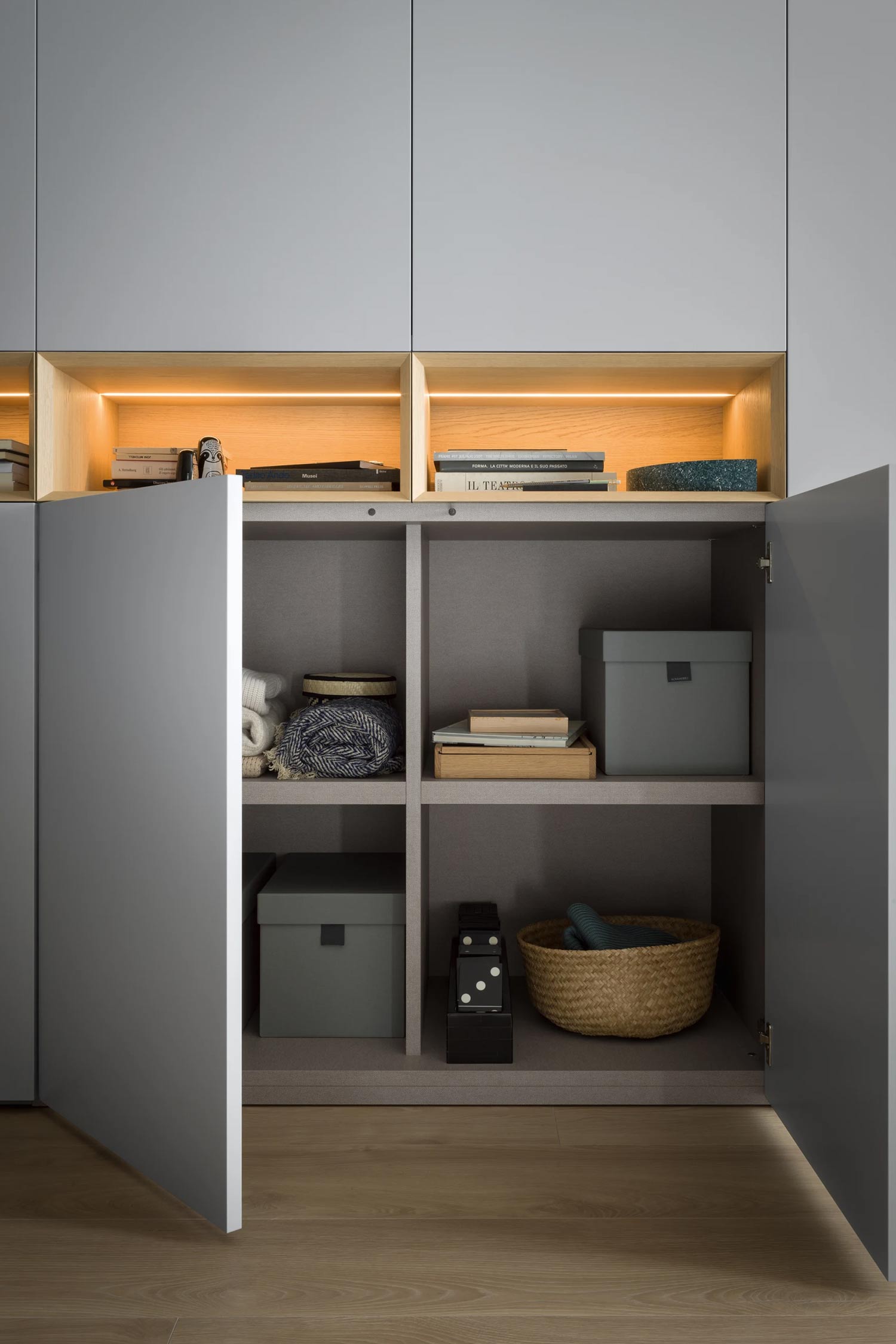 A detailed view of the open interior of a light gray wardrobe, showing organized shelves with folded items, storage boxes, and books. The top shelves are illuminated with warm lighting.