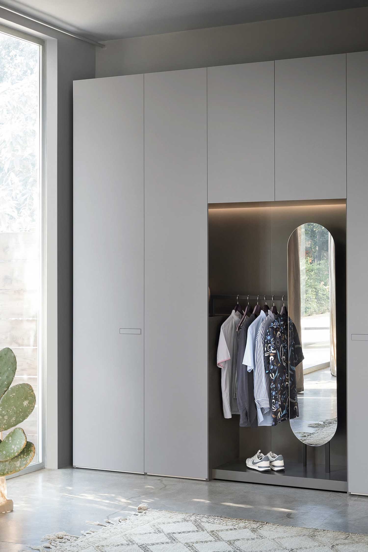 A modern, light gray hinged wardrobe with a built-in open closet section. This section has a hanging rod with shirts, a built-in shoe rack with sneakers, and a full-length, rounded mirror.