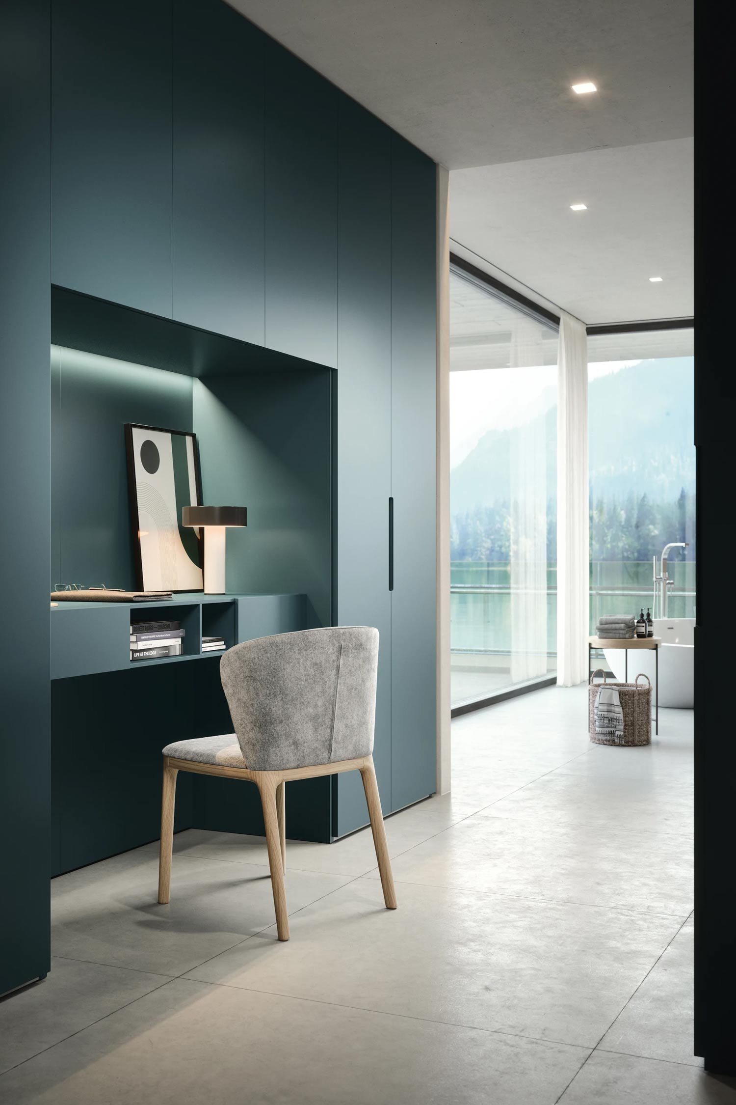 A modern home office with a built-in dark teal desk and wardrobe. The desk is recessed within the wardrobe and features a lamp and artwork. A light gray chair is pulled up to the desk, and a bright bathroom with a view of a lake and mountains is visible in the background.
