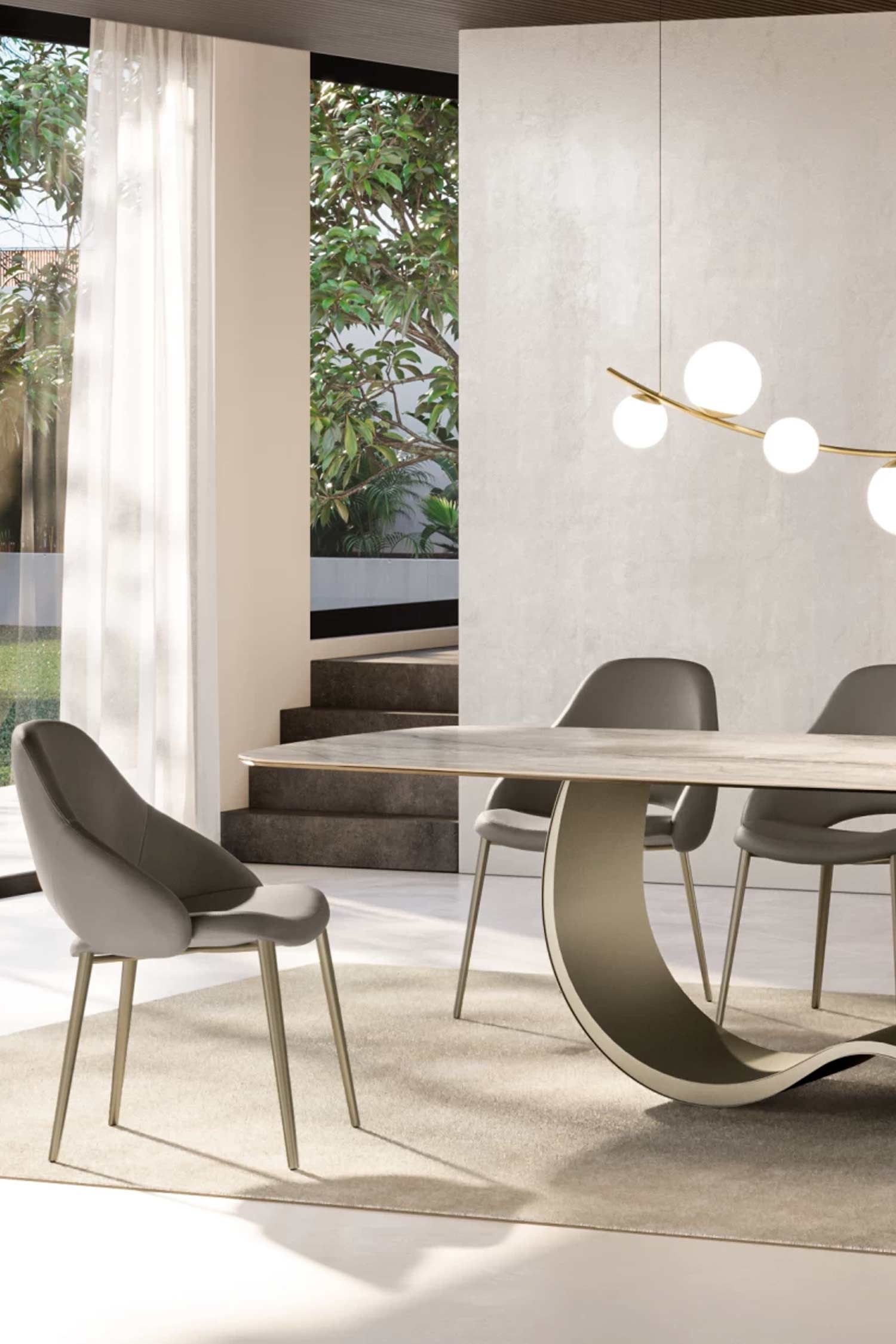 A close-up vertical view of the Alisei modern dining table with its sculptural, curved base and light-colored top. Three grey upholstered chairs are partially visible. The room has a large glass door leading to a lush green outdoor area, with sheer white curtains providing natural light. A simple brass and white globe chandelier hangs above.