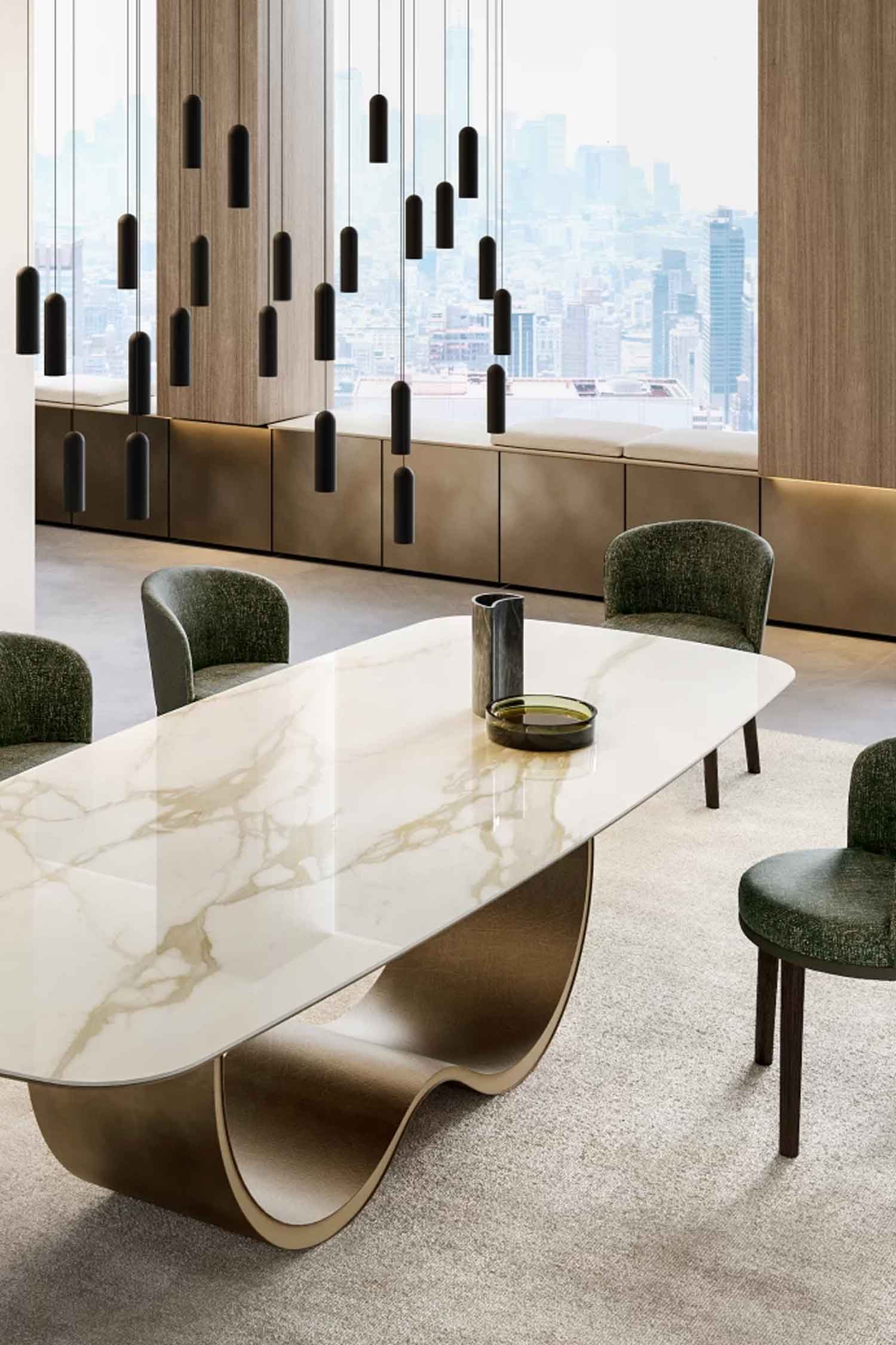 A vertical close-up of the Alisei luxury dining table with its polished white marble top featuring gold veining and a wavy, sculptural bronze base. Four dark green textured dining chairs are placed around the table, over which several sleek black cylindrical pendant lights hang. A large window offers a city view in the background.