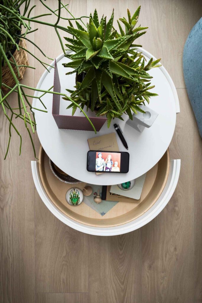 Overhead close-up of a modern round white nightstand with a partially open wood drawer, showing an aloe plant, a cell phone displaying a photo, a pen, and a handwritten note.