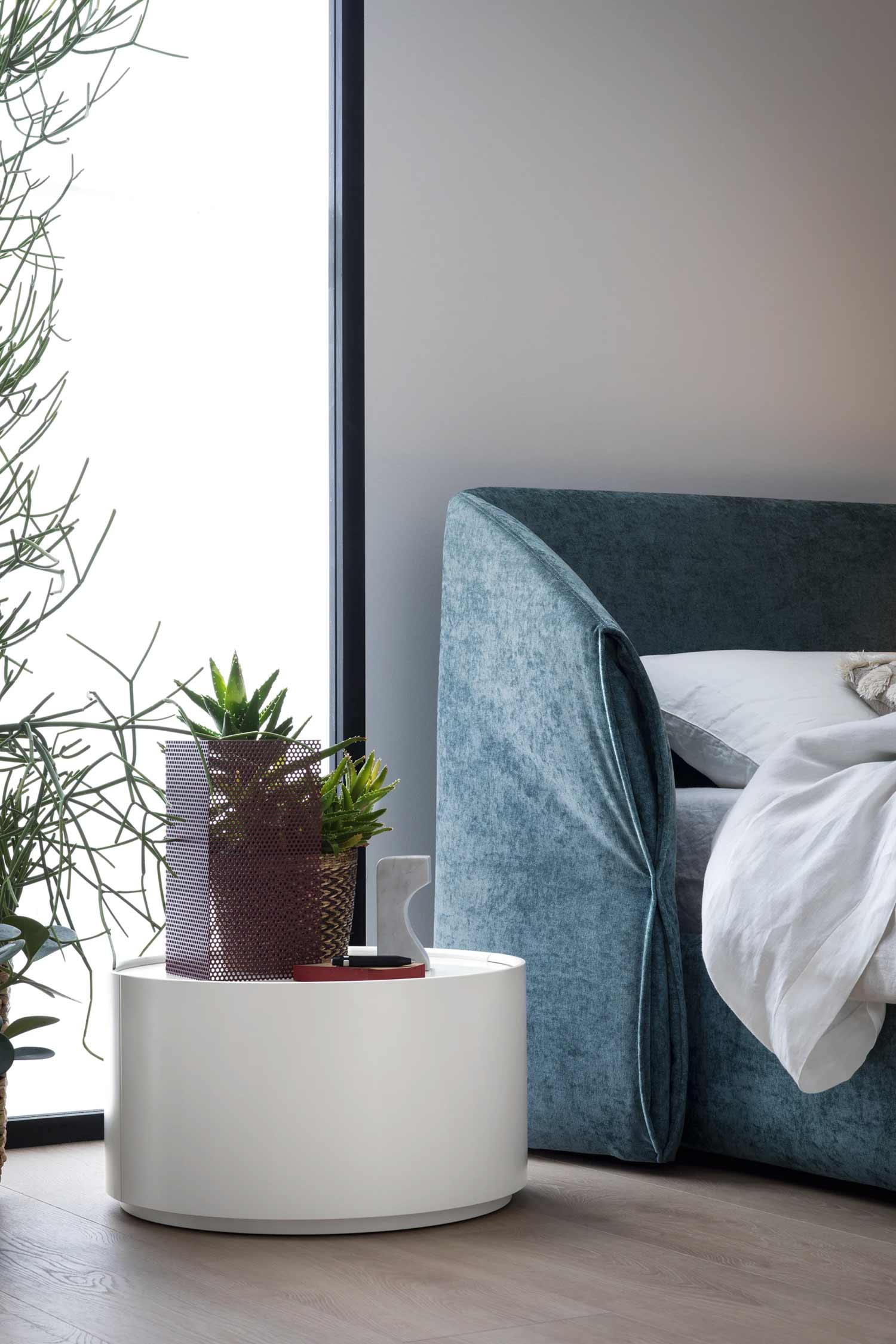 Close-up of a modern white circular nightstand with a small potted plant and a metal accent piece, next to a bed with a high, rounded, textured teal velvet headboard.