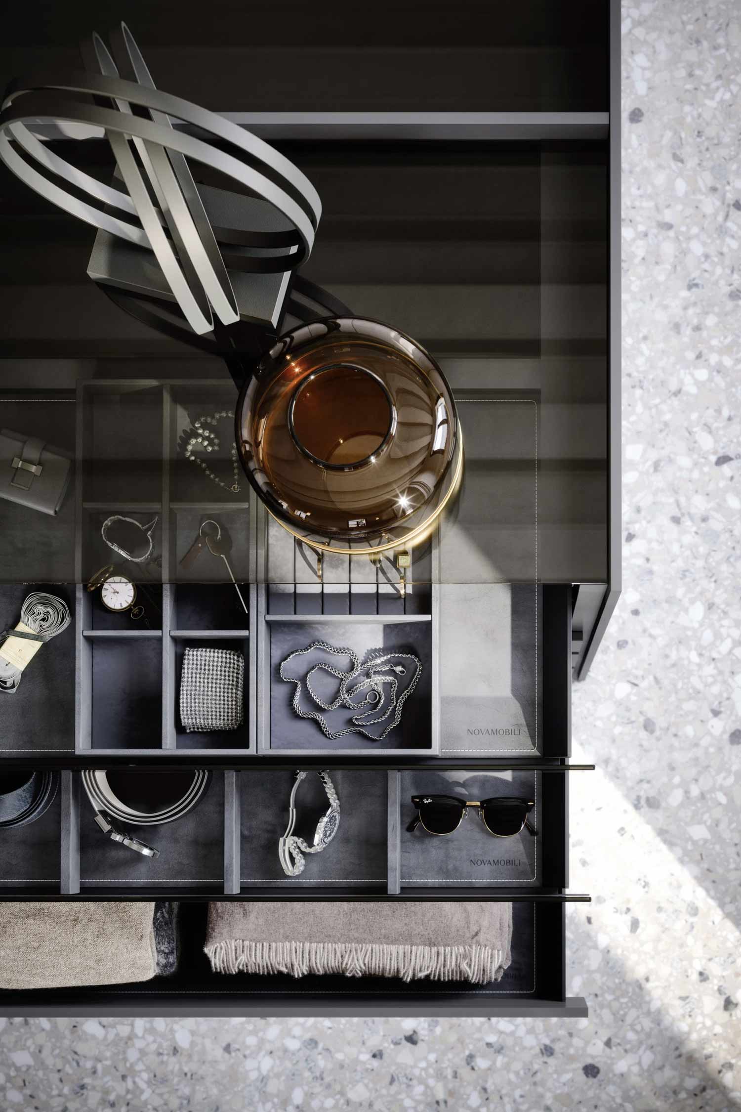 A high-angle view looking down into an open drawer of a closet island. The drawer is organized with various accessories, including a watch, a silver chain, sunglasses, and a belt. The accessories are separated by gray dividers. A circular, amber-colored glass object and a sculpture made of black and white bands are also visible. The floor is a light-colored terrazzo.