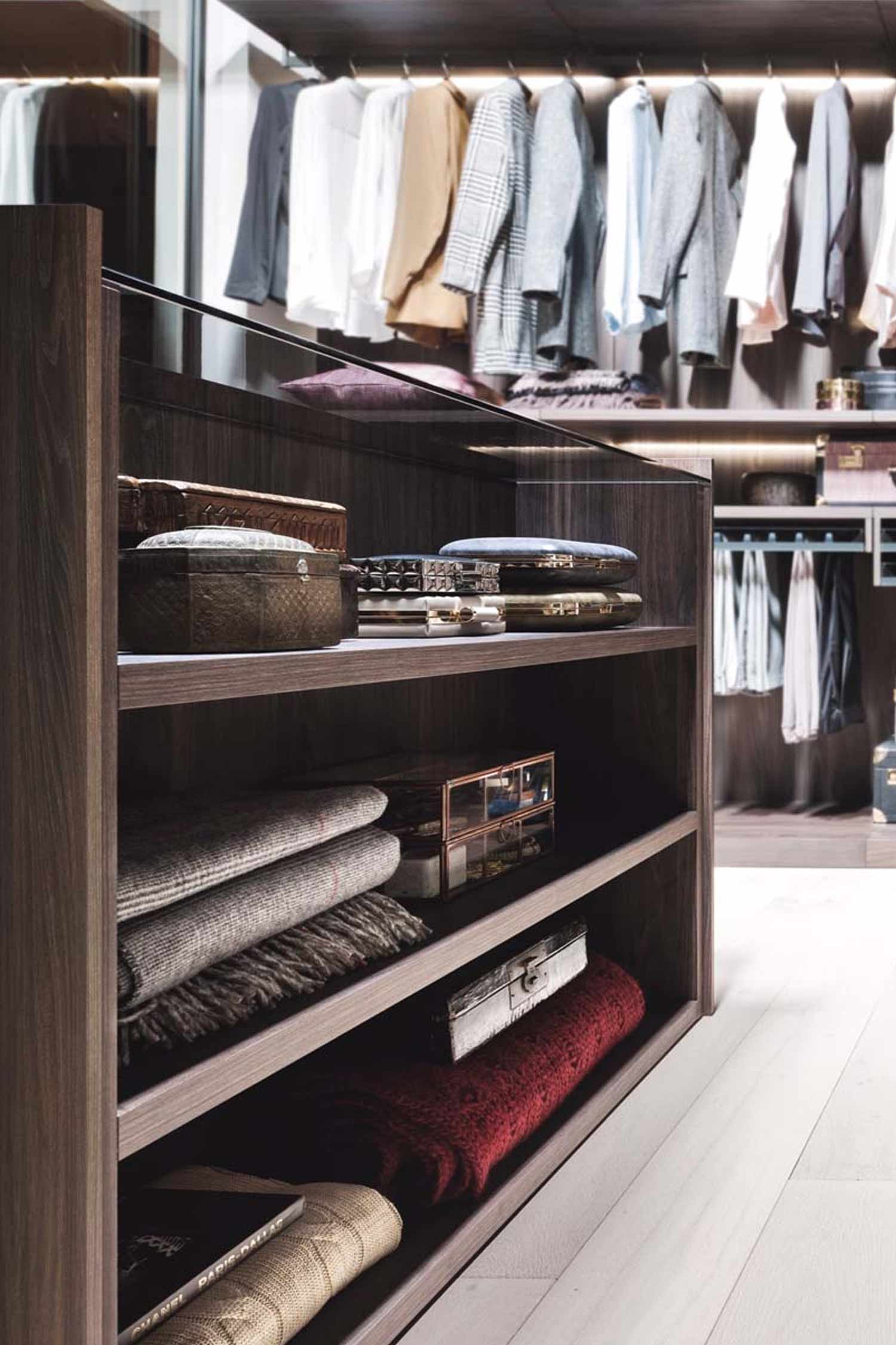 A close-up, angled view of a dark wood walk-in closet with shelves filled with folded blankets, a book, and two decorative boxes. The top shelf has a glass front. In the background, clothes are visible hanging on a rack.