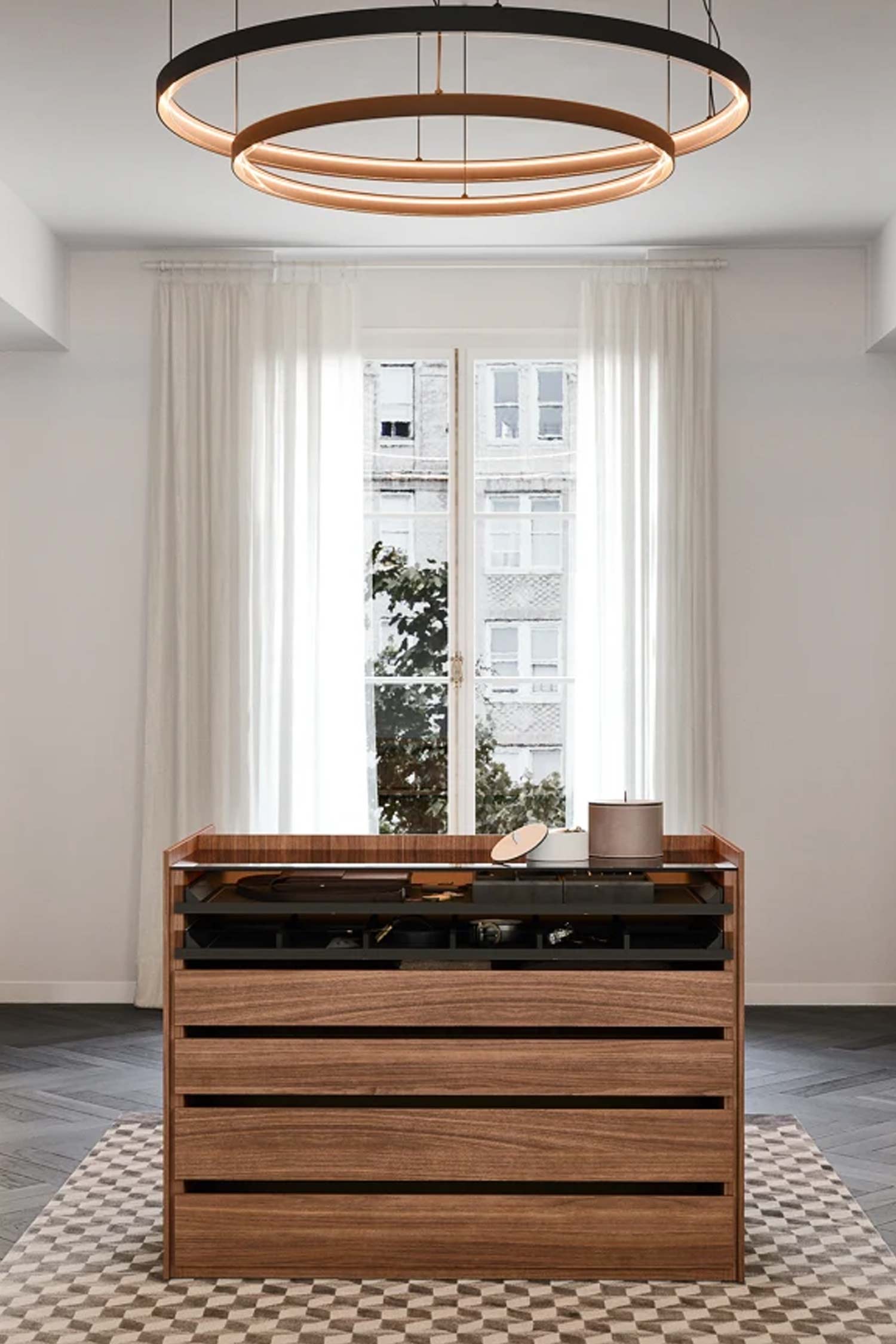 A front-facing view of a wooden walk-in closet island with a glass top. The island has four drawers, and the top is open, revealing a black divided tray for accessories. The island sits on a checkered rug, centered in front of a window with sheer white curtains. Two circular pendant lights hang from the ceiling.
